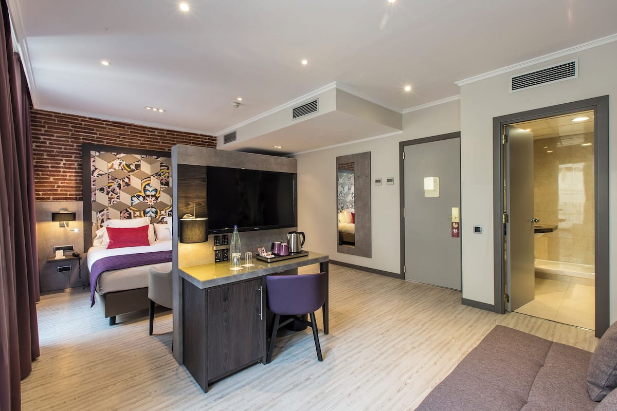 Moderni hotellihuone, jossa on sänky, työpöytä, tuoli, televisio ja suihkutila.