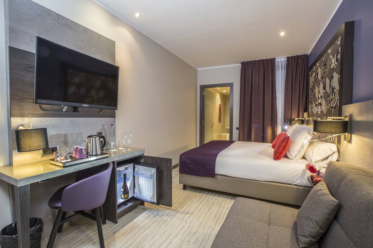 Moderni hotellihuone, jossa on sänky, työpöytä tietokoneella, televisio ja minijääkaappi.