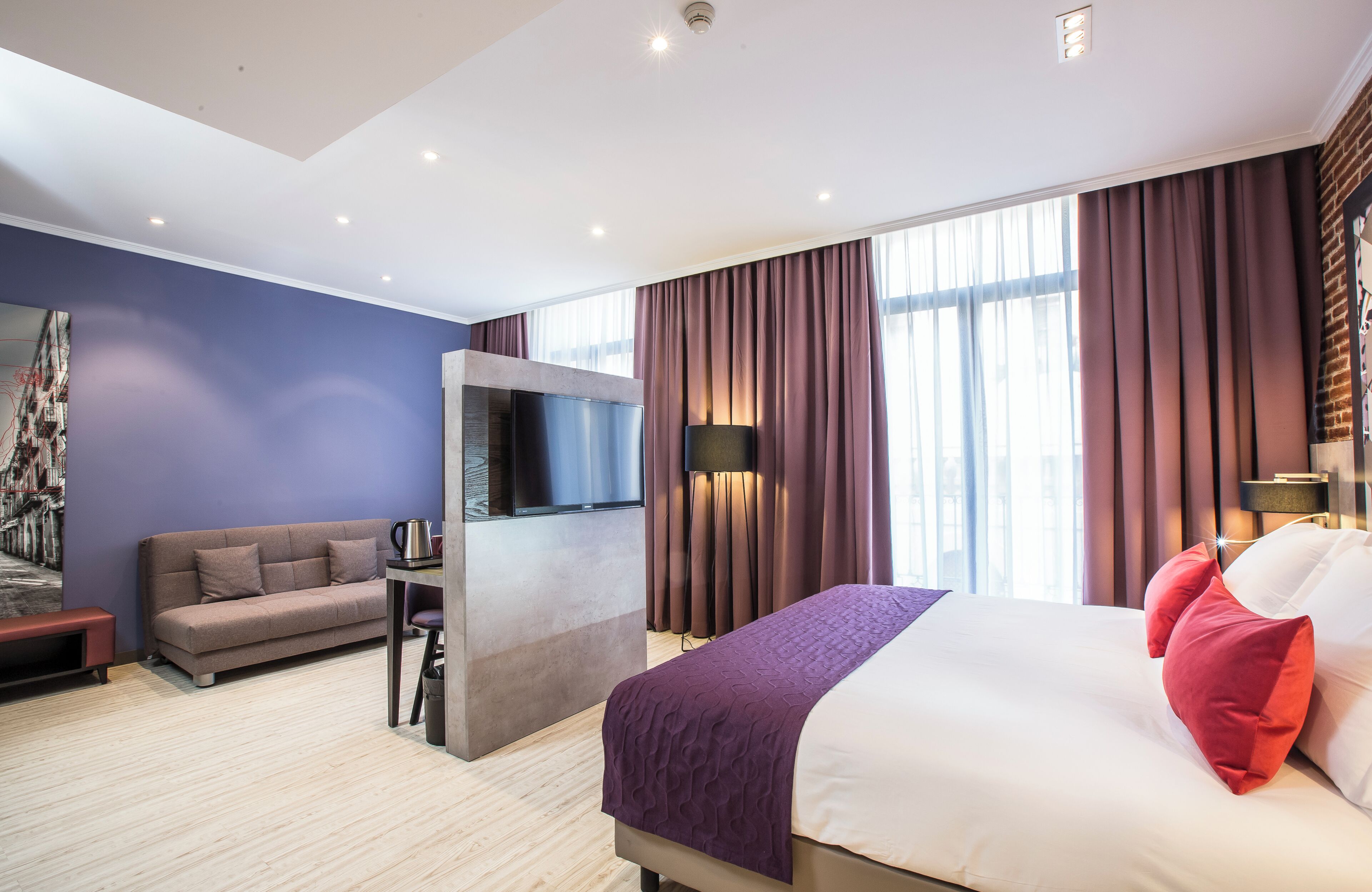 Moderni hotellihuone, jossa on sänky, sohva, televisio ja suuri ikkuna, jossa on verhot.