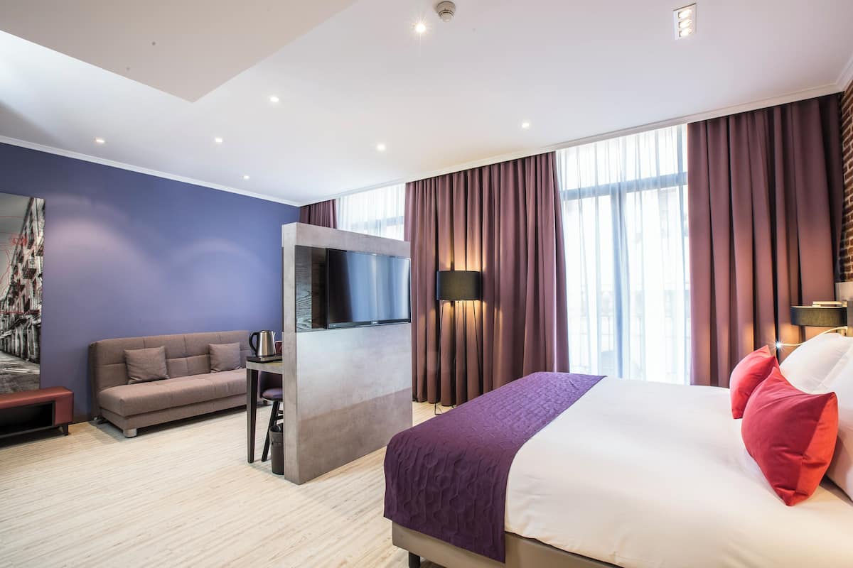 Moderni hotellihuone, jossa on sänky, sohva, televisio ja suuri ikkuna, jossa on verhot.