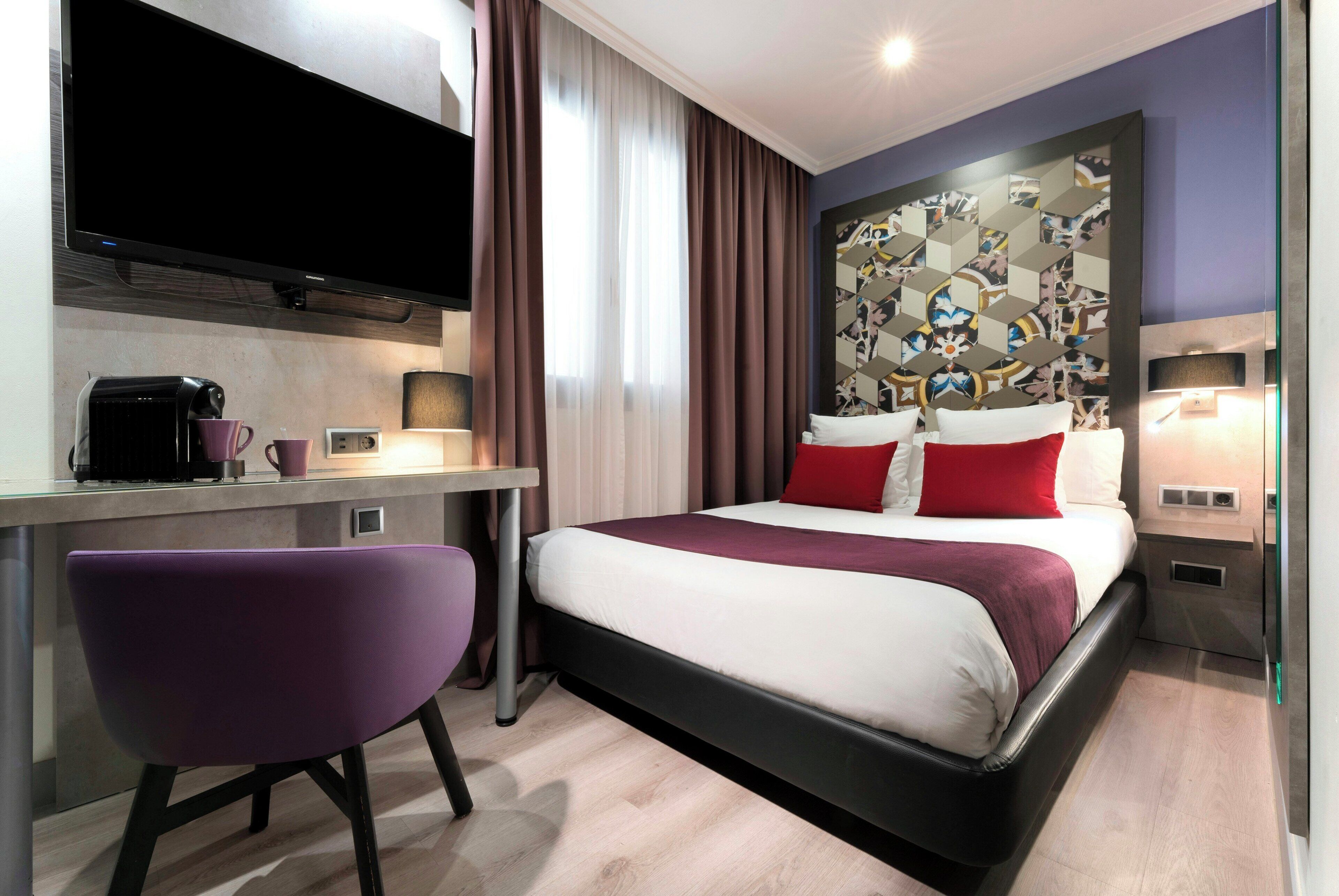 A hotel room with a bed, a desk, a chair, a flat-screen TV, and a wall with a geometric pattern.