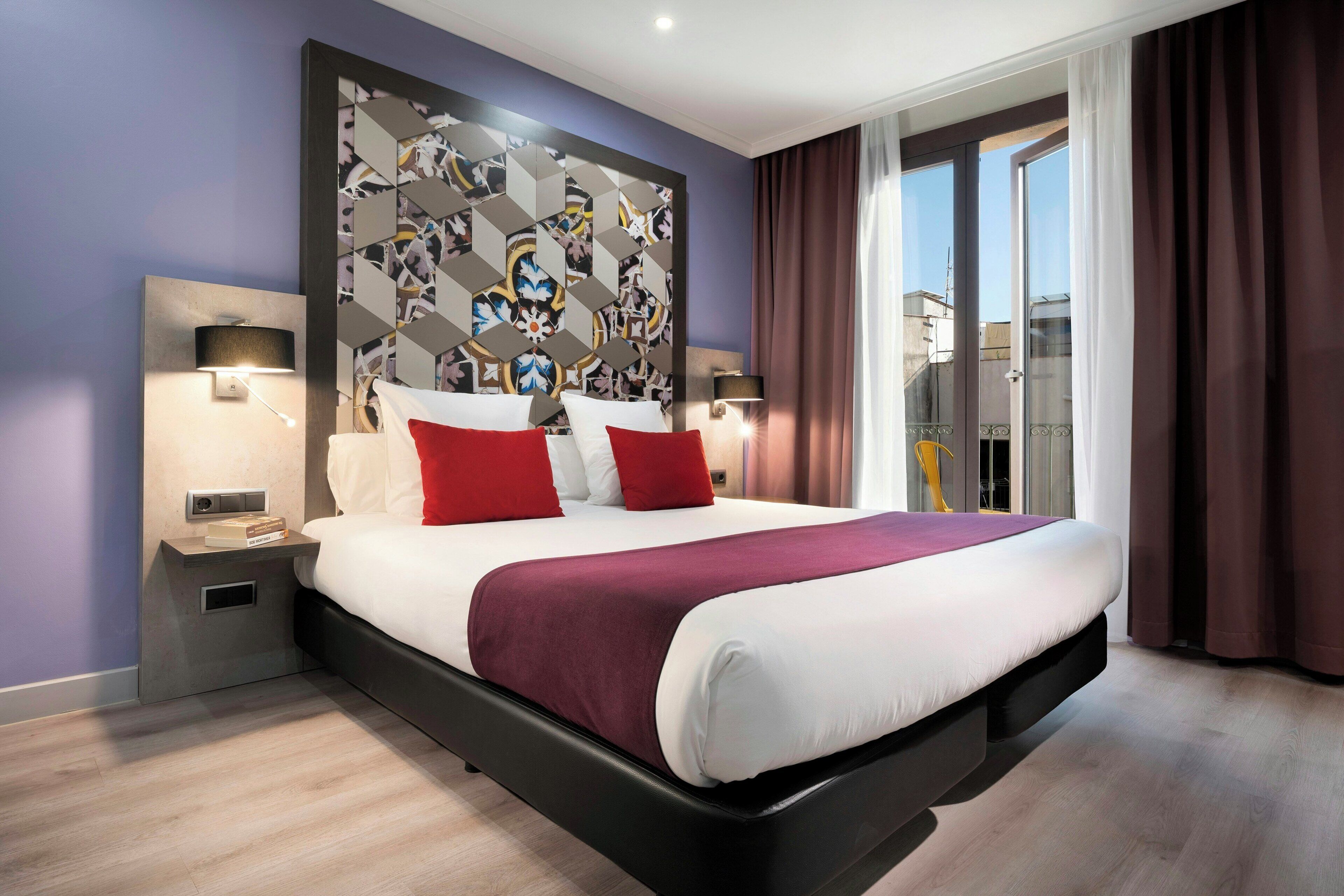 A modern hotel room with a large bed, two bedside tables, a decorative wall panel, and a view of the outside.