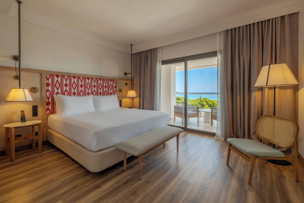 Habitación de hotel con una cama grande, un banco, una silla, una mesa y vistas al exterior.