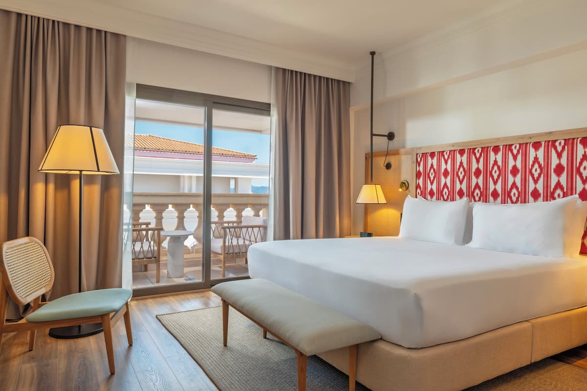 Habitación de hotel con una cama grande, una silla, un banco, una lámpara y vistas al exterior.