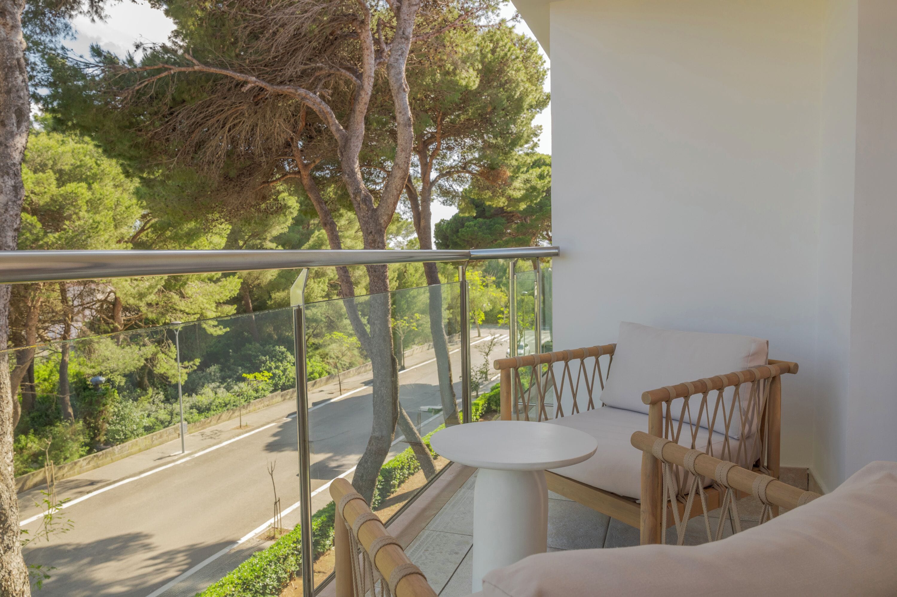 A balcony with a view of a road and trees, a white table, and a wooden chair.