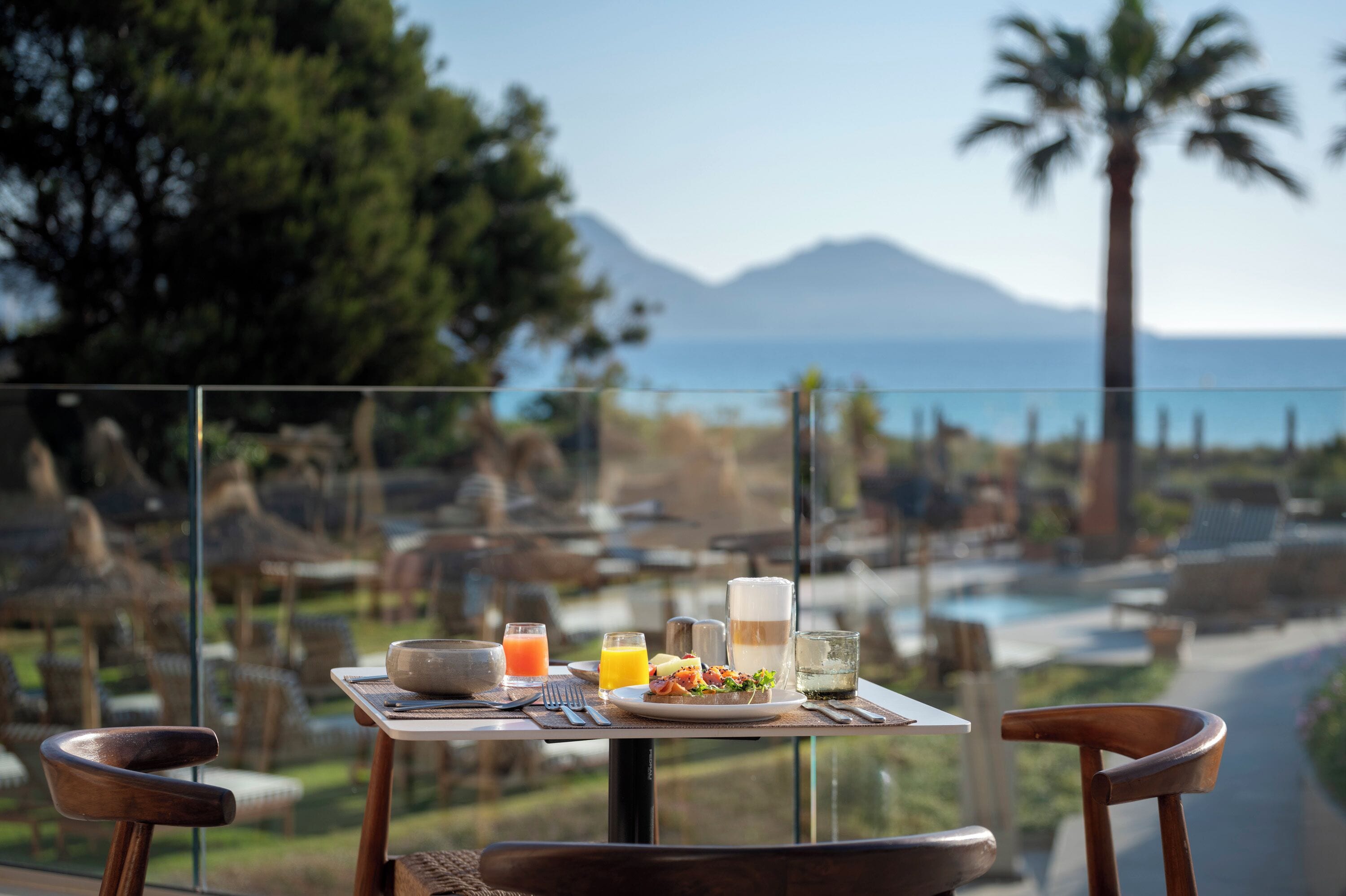 A dining table with breakfast items and a glass of orange juice, overlooking a scenic view with a pool and mountains.