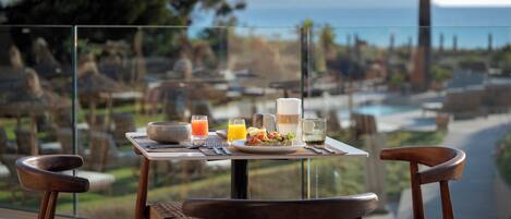 A dining table with breakfast items and a glass of orange juice, overlooking a scenic view with a pool and mountains.