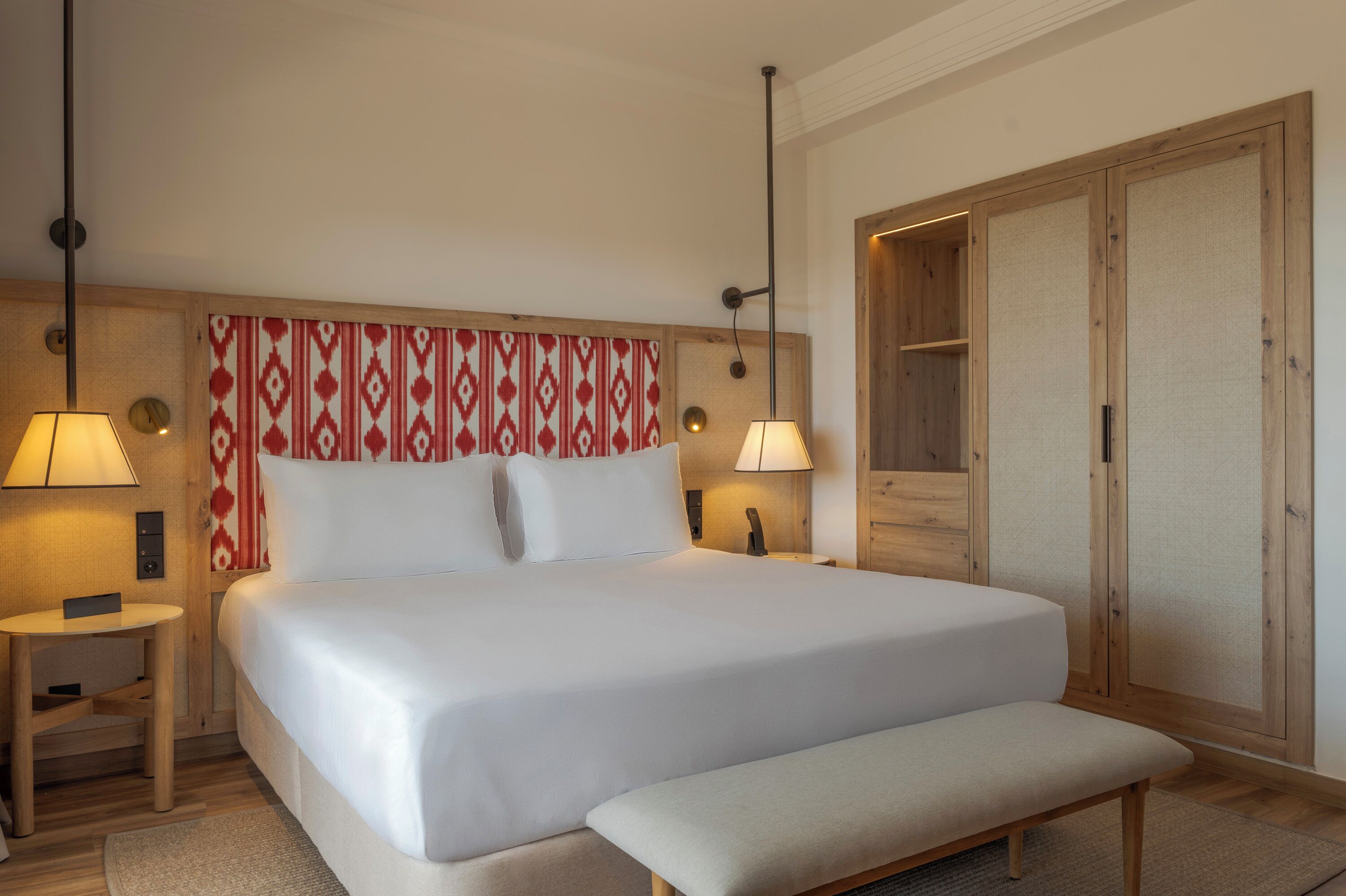 Una habitación de hotel con una cama grande, lámparas de noche, un armario de madera y un banco.
