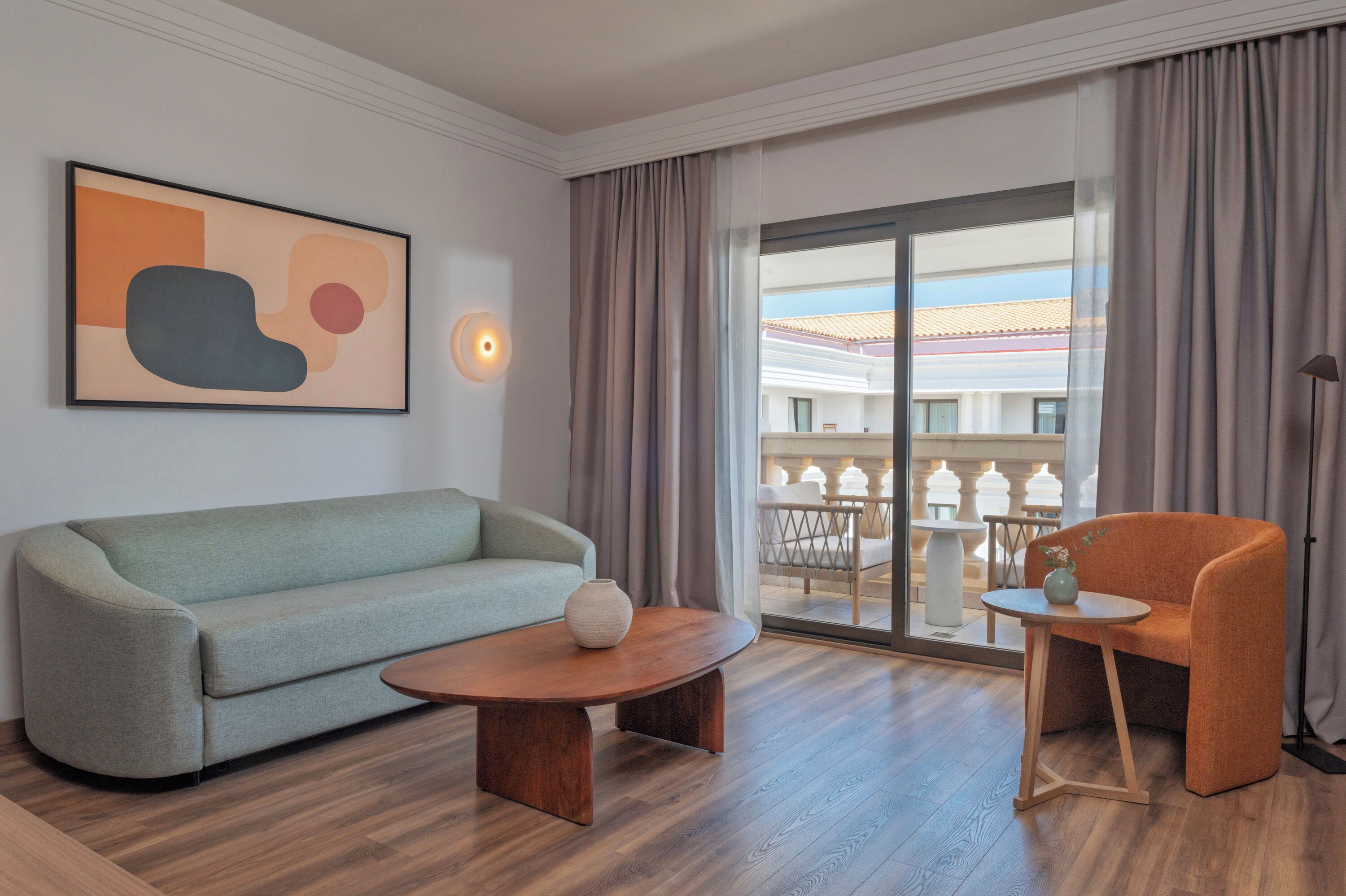 Ein modernes Hotelzimmer mit einer Couch, einem kleinen Tisch, einem Sessel, einem großen Fenster mit Vorhängen und einer Wanddekoration.