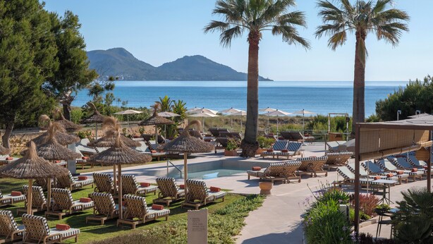Un resort con piscina, tumbonas, palmeras y vistas al mar y a las montañas.