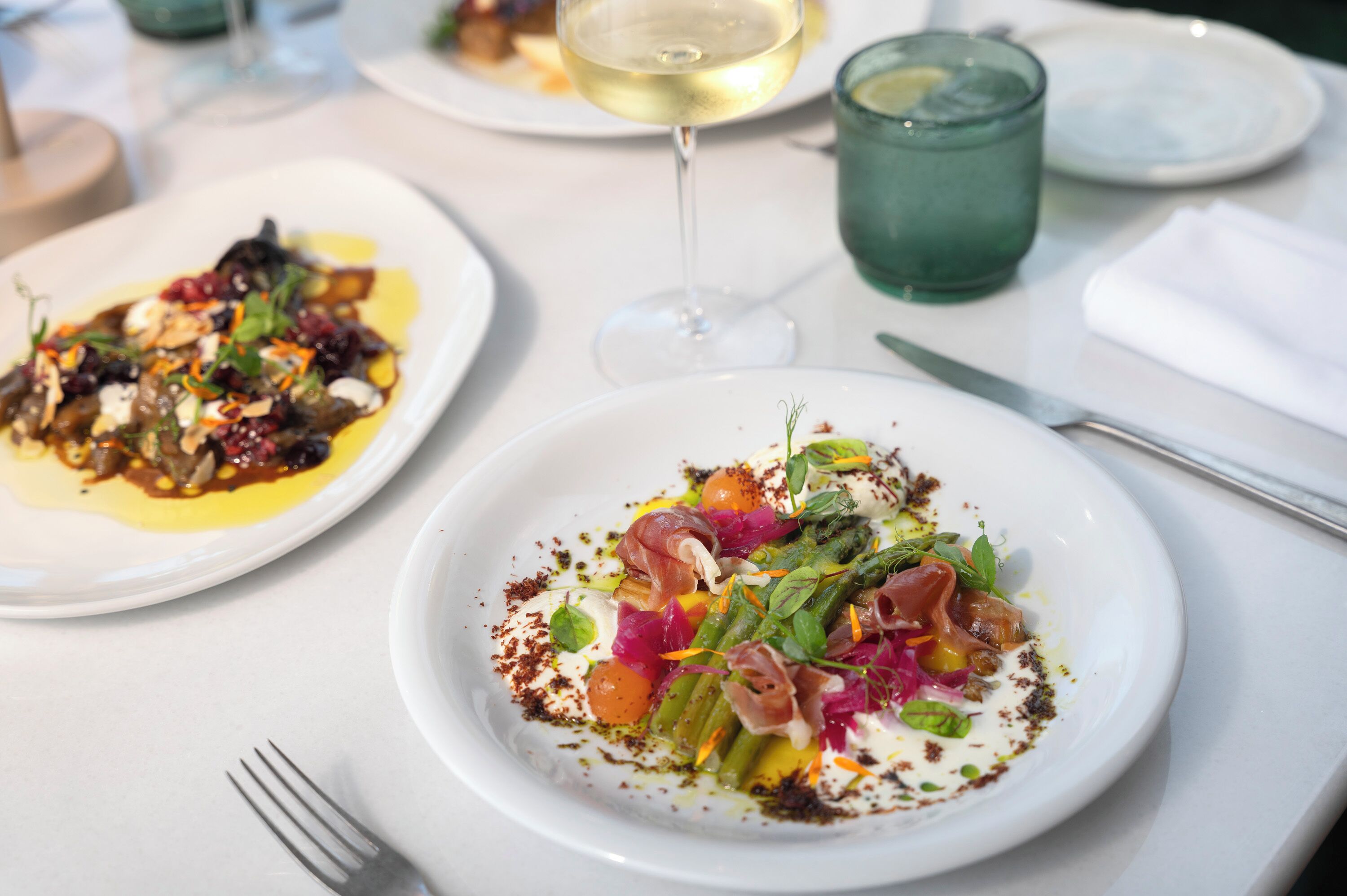 A dining table with plates of food, a glass of white wine, and a green glass.