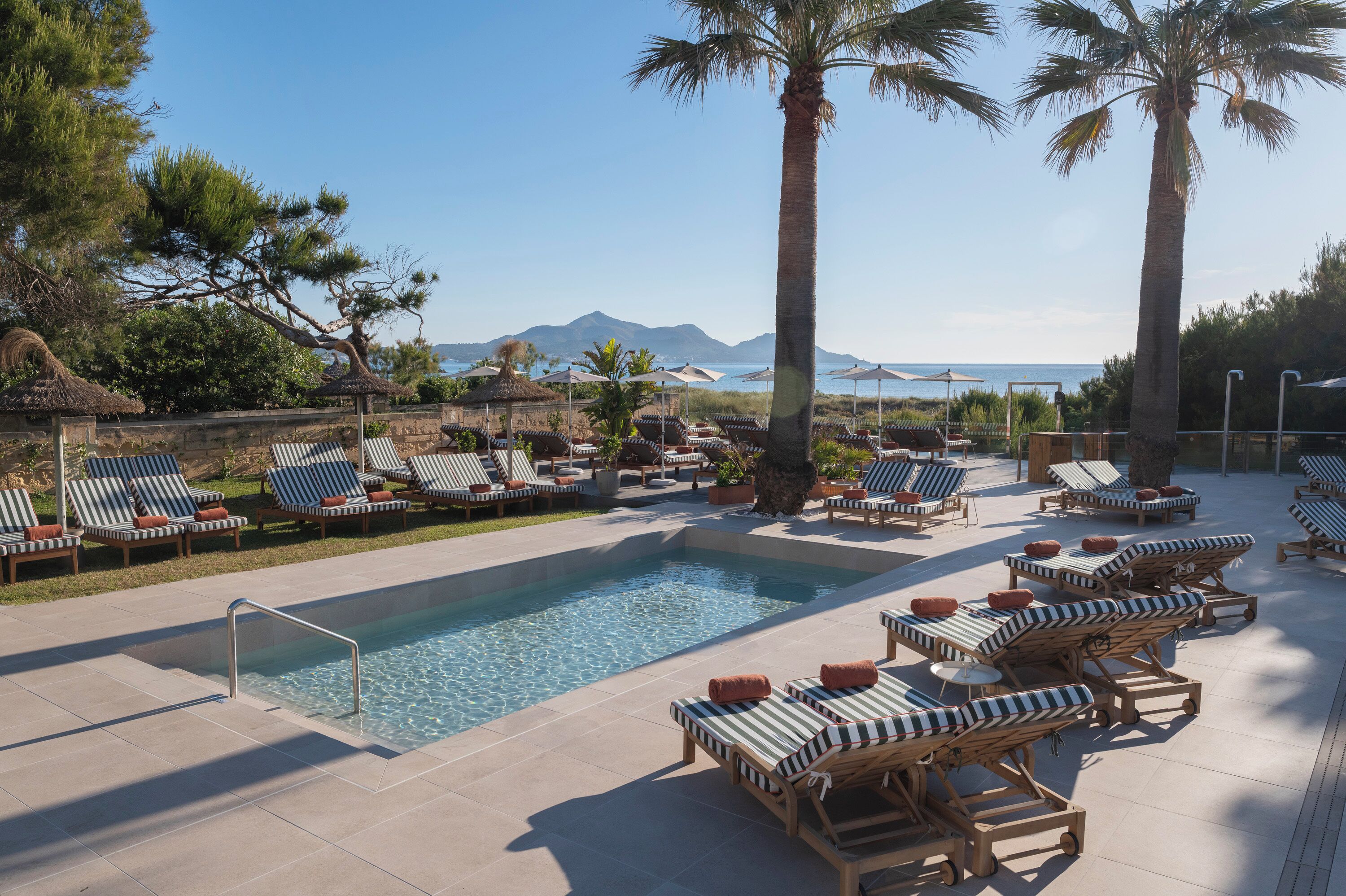 A poolside area with lounge chairs, a swimming pool, and a view of the sea and mountains.