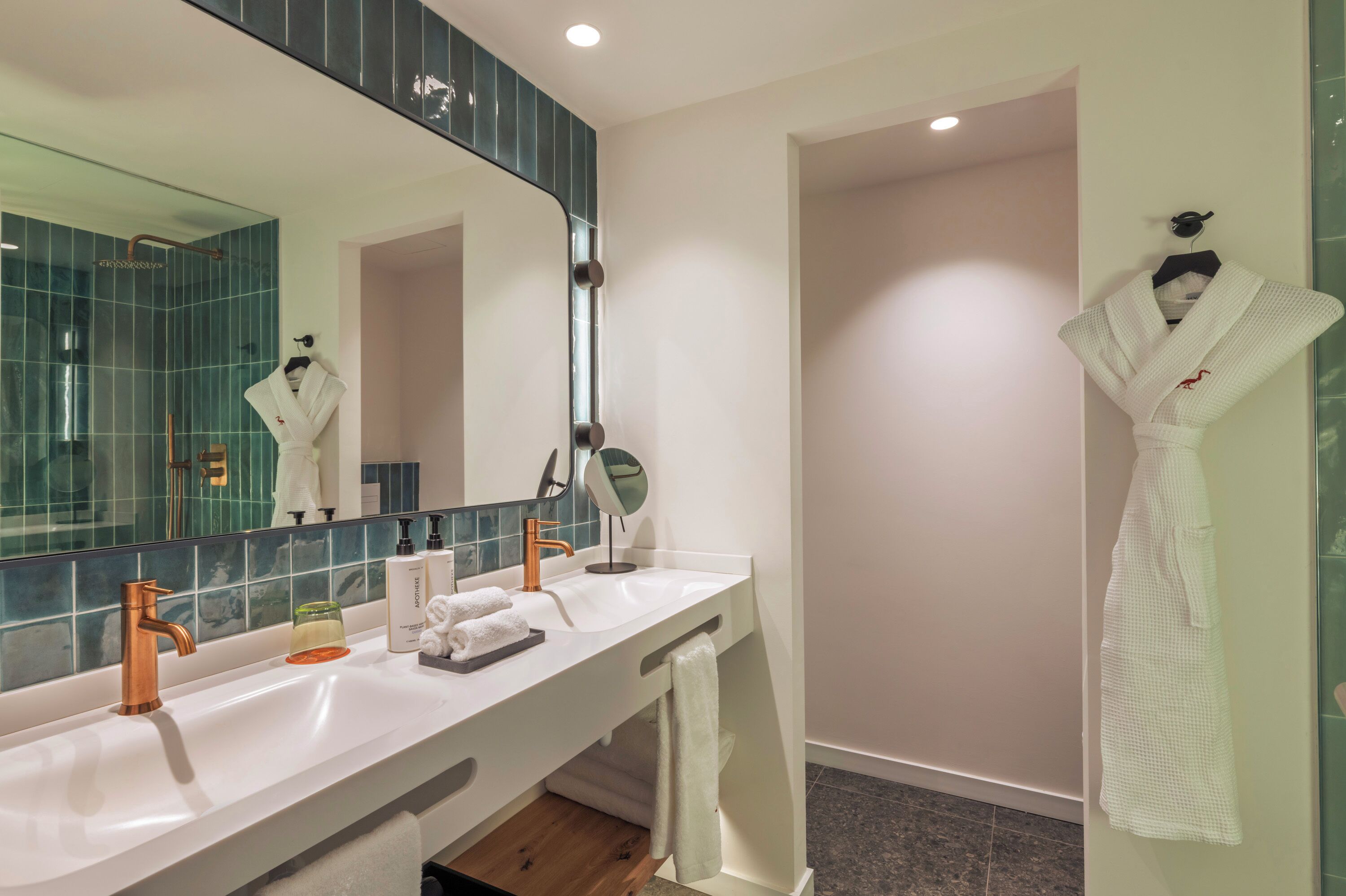 A modern bathroom with a large mirror, two sinks, a towel rack, and a robe hanging on the wall.