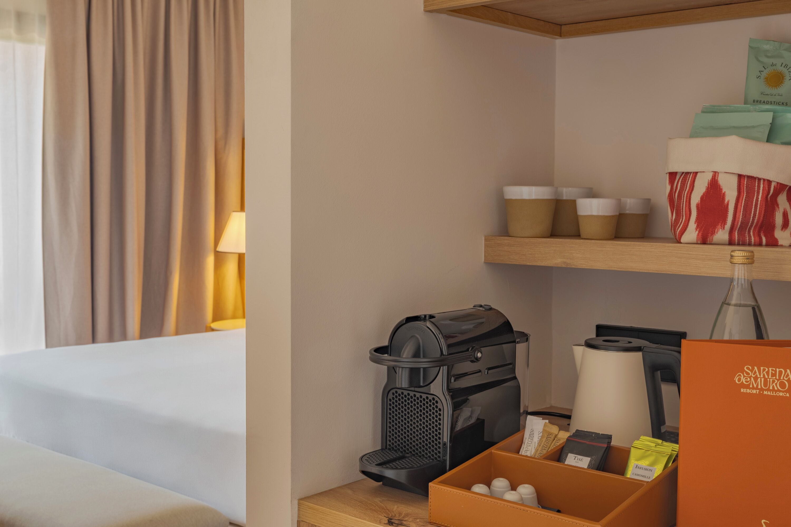 Habitación de hotel con cama, cafetera, una botella y una caja de palitos de pan.