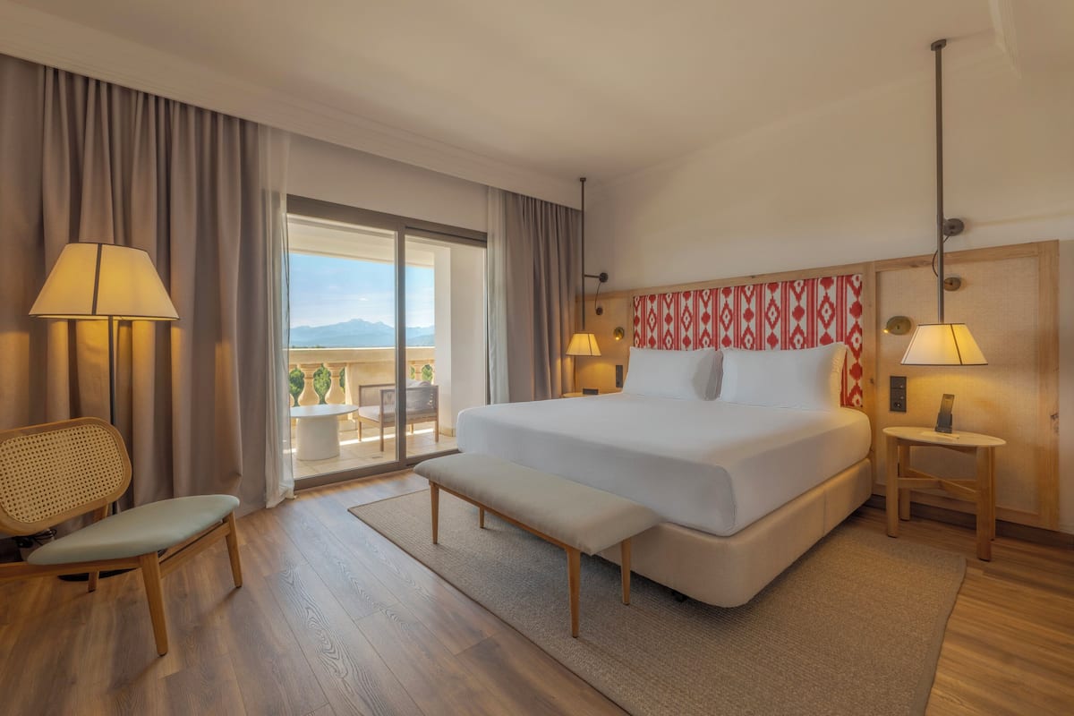 Una habitación de hotel con una cama grande, dos mesitas de noche con lámparas, un banco, una silla y vistas amplias por una ventana grande.