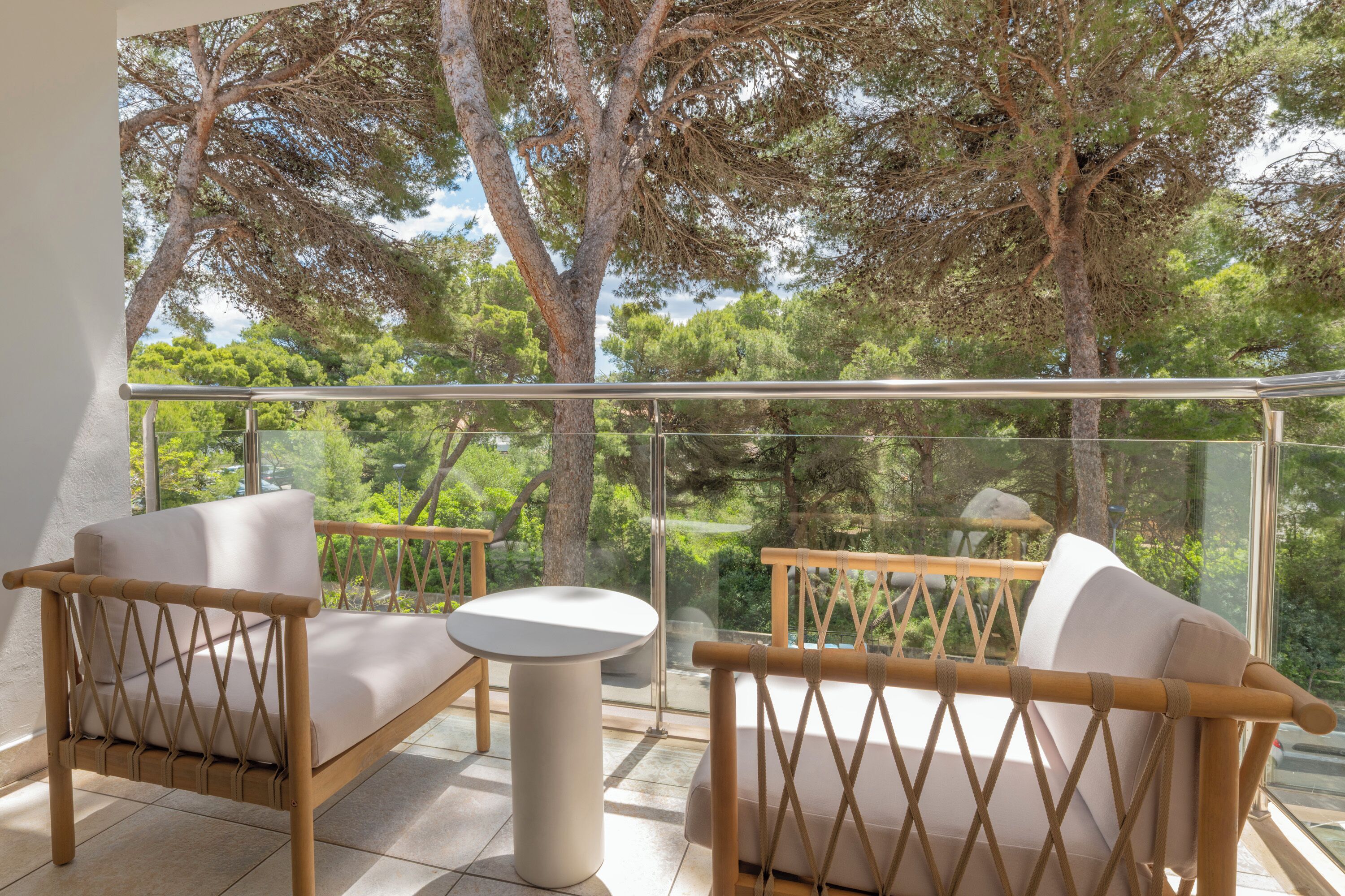 A balcony with a glass railing, two wicker chairs, a small round table, and a view of trees.