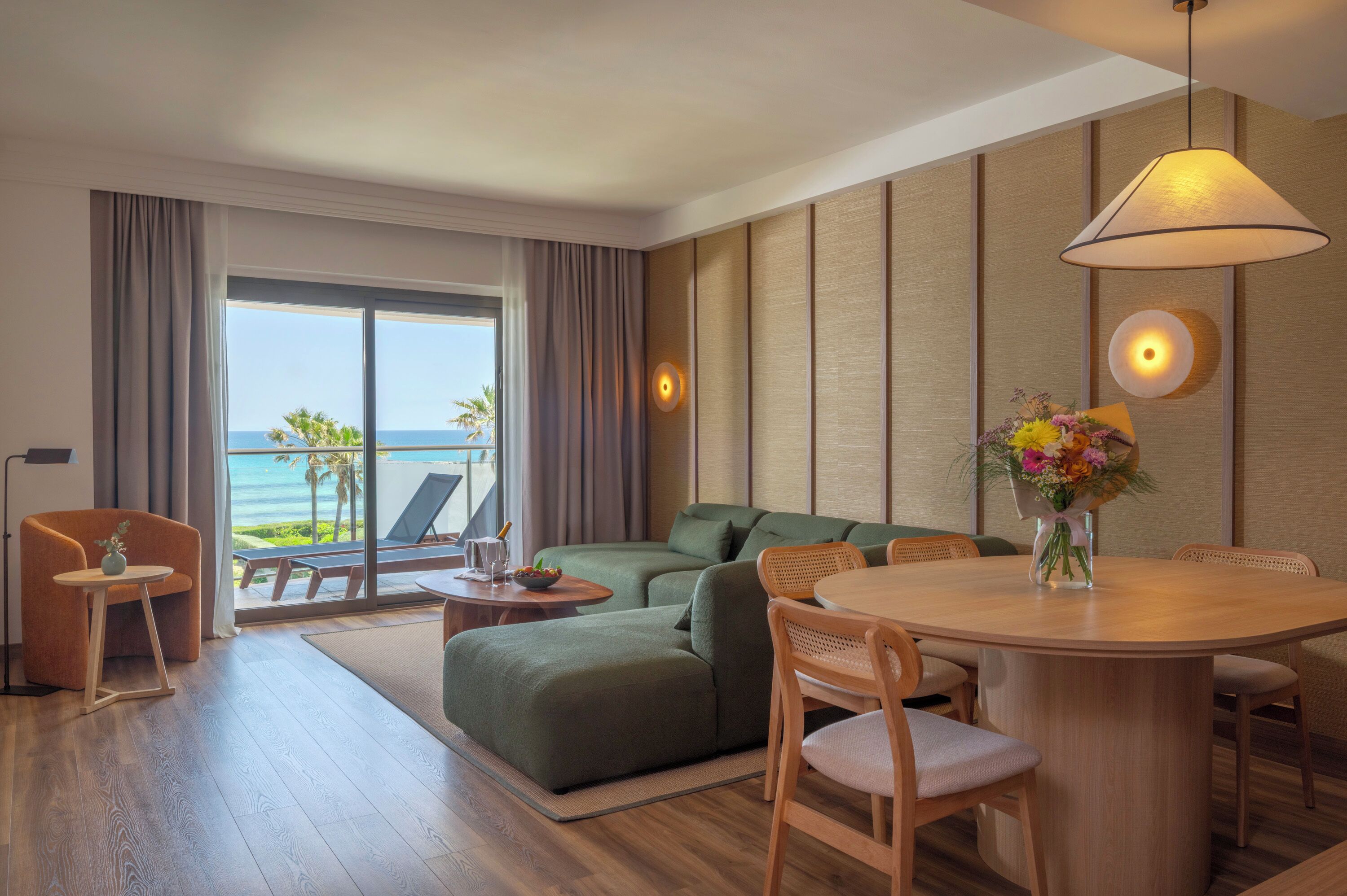 A modern hotel room with a large sliding glass door to the beach, a green sofa, a wooden dining table, and a small side table with a vase of flowers.
