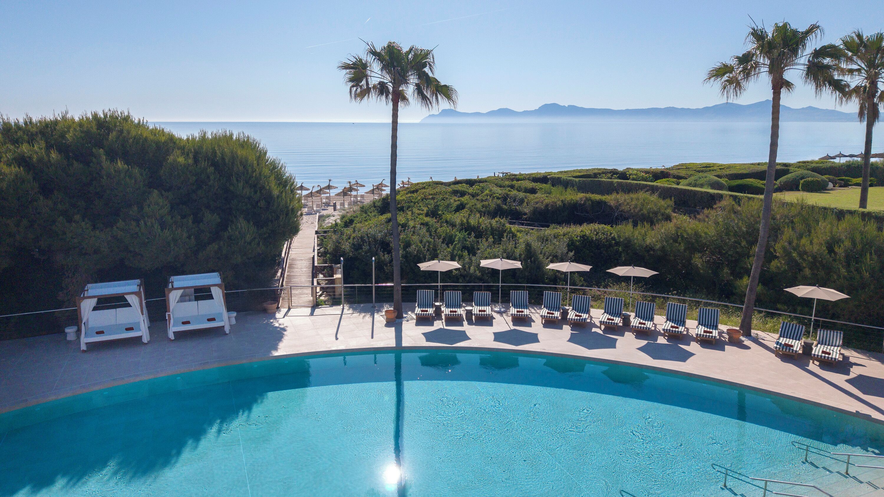 A pool with lounge chairs and umbrellas, surrounded by greenery and a view of the sea.