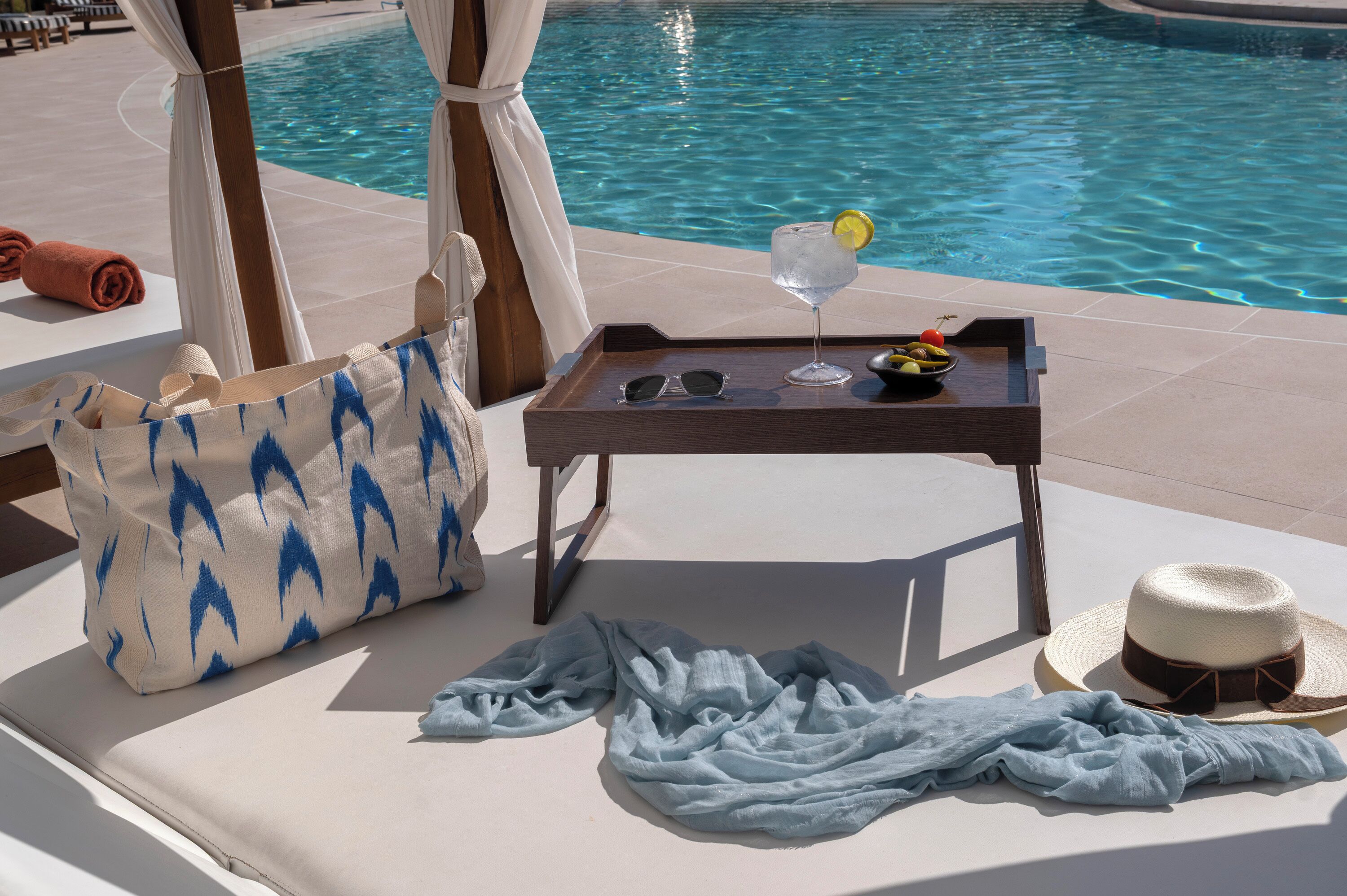 Zona de descanso junto a la piscina con una tumbona, una mesa con una bebida, gafas de sol y un sombrero.