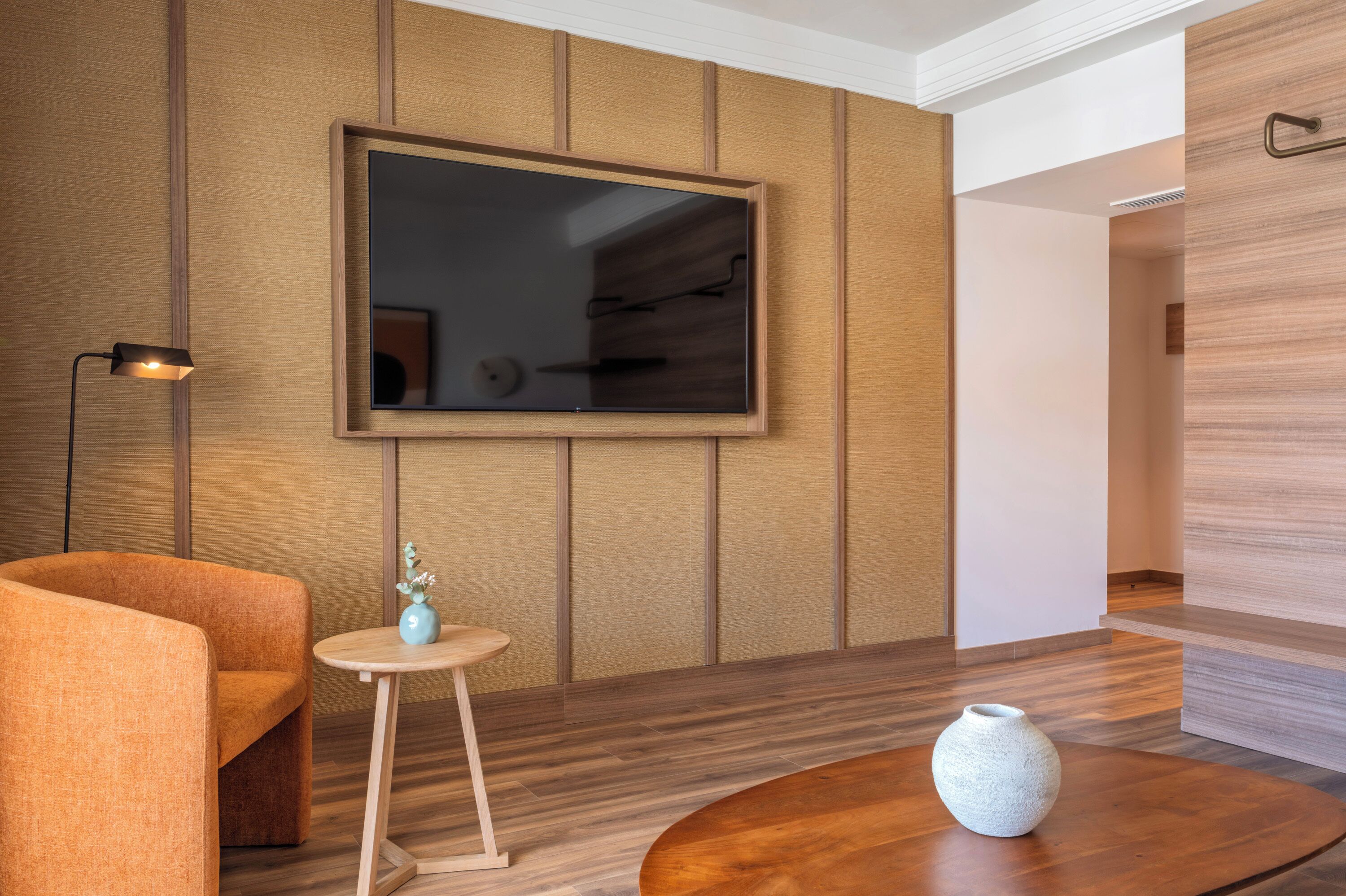 Une chambre moderne dotée d’un grand téléviseur à écran plat fixé au mur, d’un plancher en bois, d’une petite table ronde ornée d’un vase et d’une seule chaise orange.