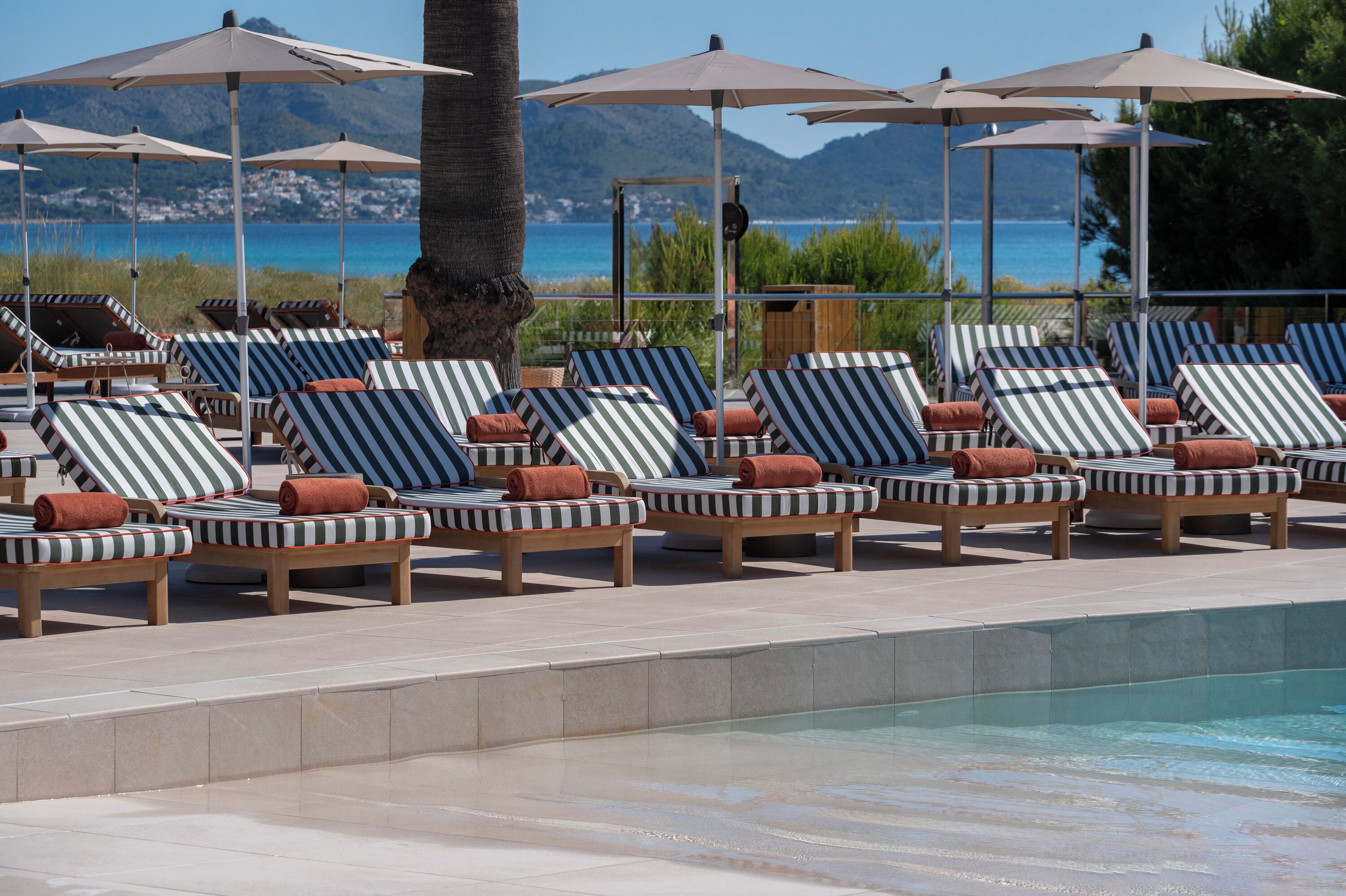 A poolside area with lounge chairs and umbrellas, a clear view of the sea, and a mountainous backdrop.