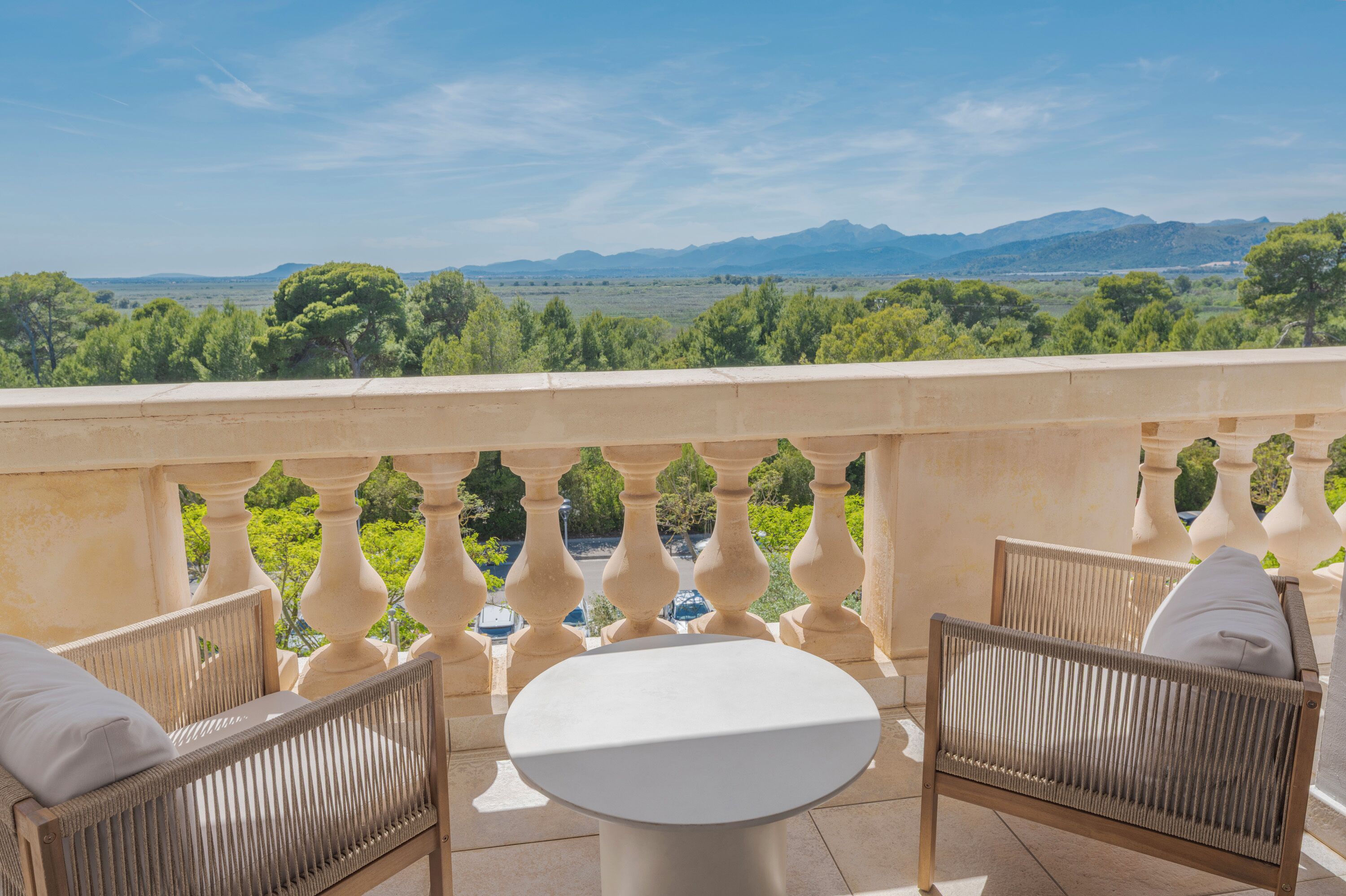 A balcony with wicker furniture, a round table, and a view of distant mountains and greenery.
