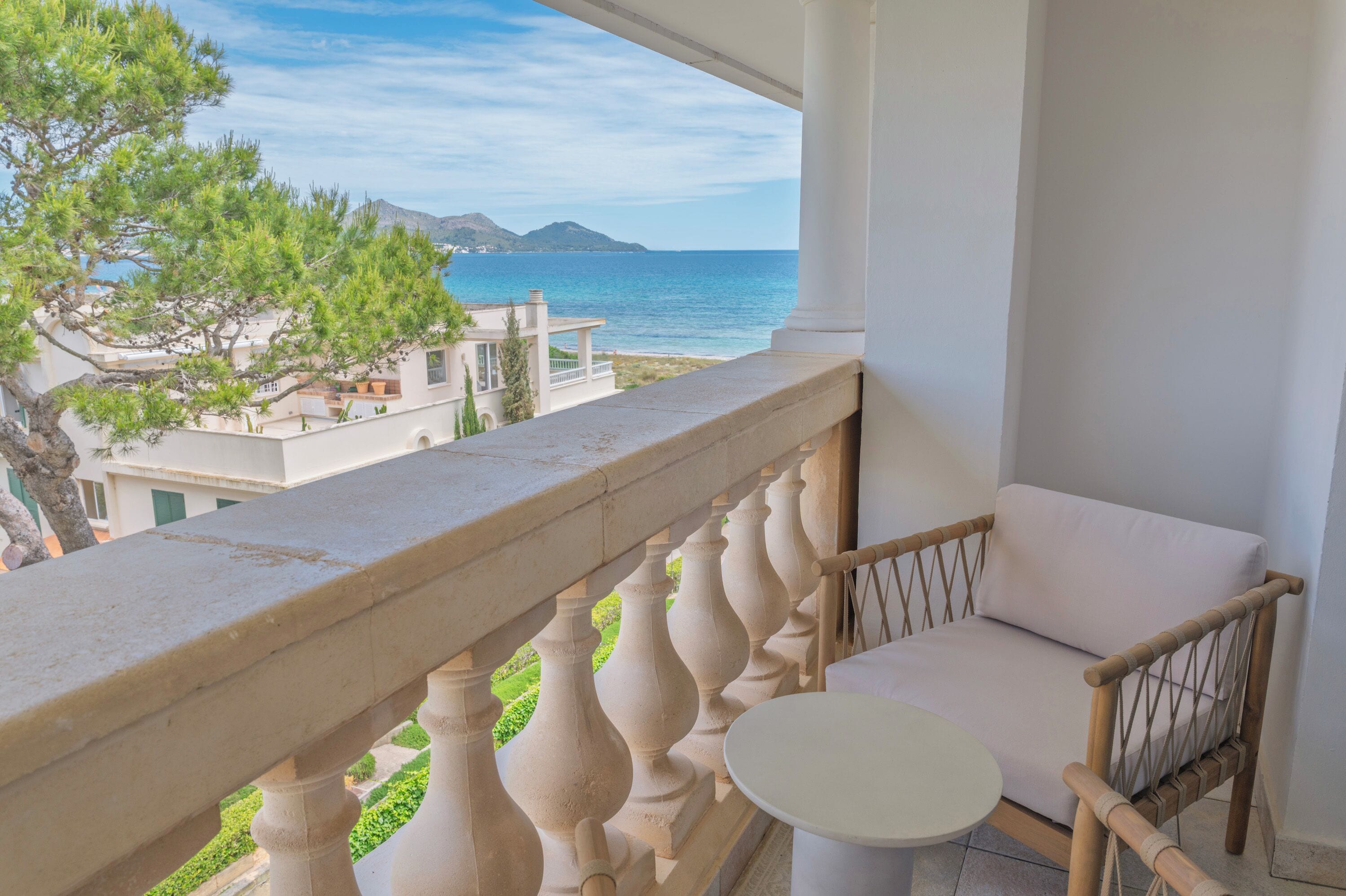 A balcony with a white cushioned chair and a small table, overlooking a coastal view with buildings and greenery.