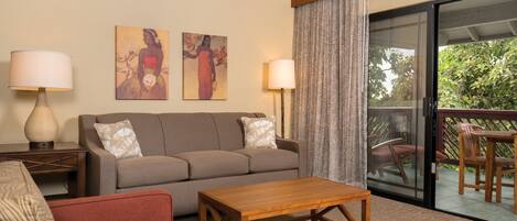 Une chambre d’hôtel avec un canapé, une table basse en bois, un fauteuil rouge, deux tableaux au mur, une lampe et une porte-fenêtre coulissante donnant sur un espace extérieur avec une table et des chaises.