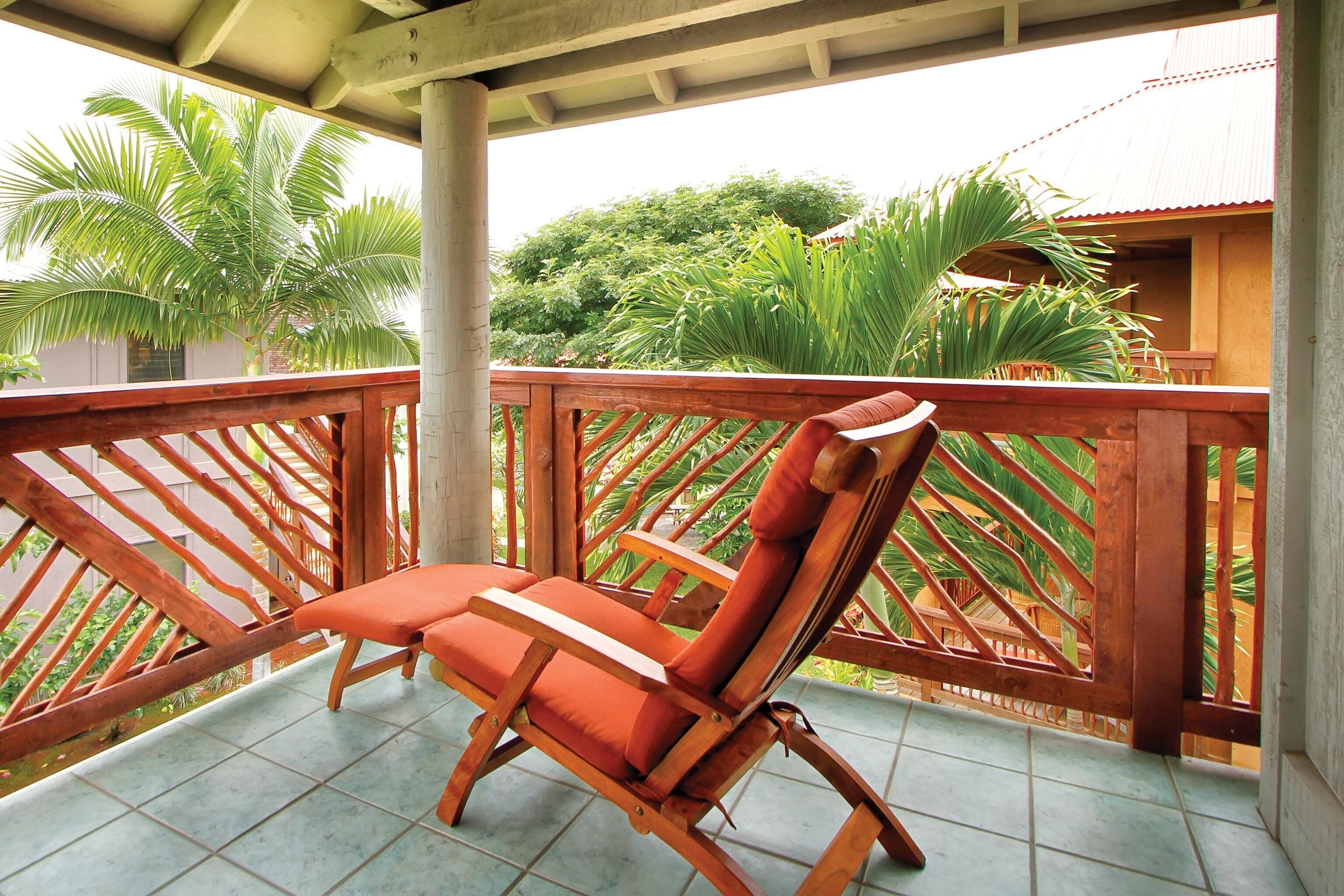 A wooden chair with a cushion on a balcony with a tiled floor, surrounded by a wooden railing and greenery.