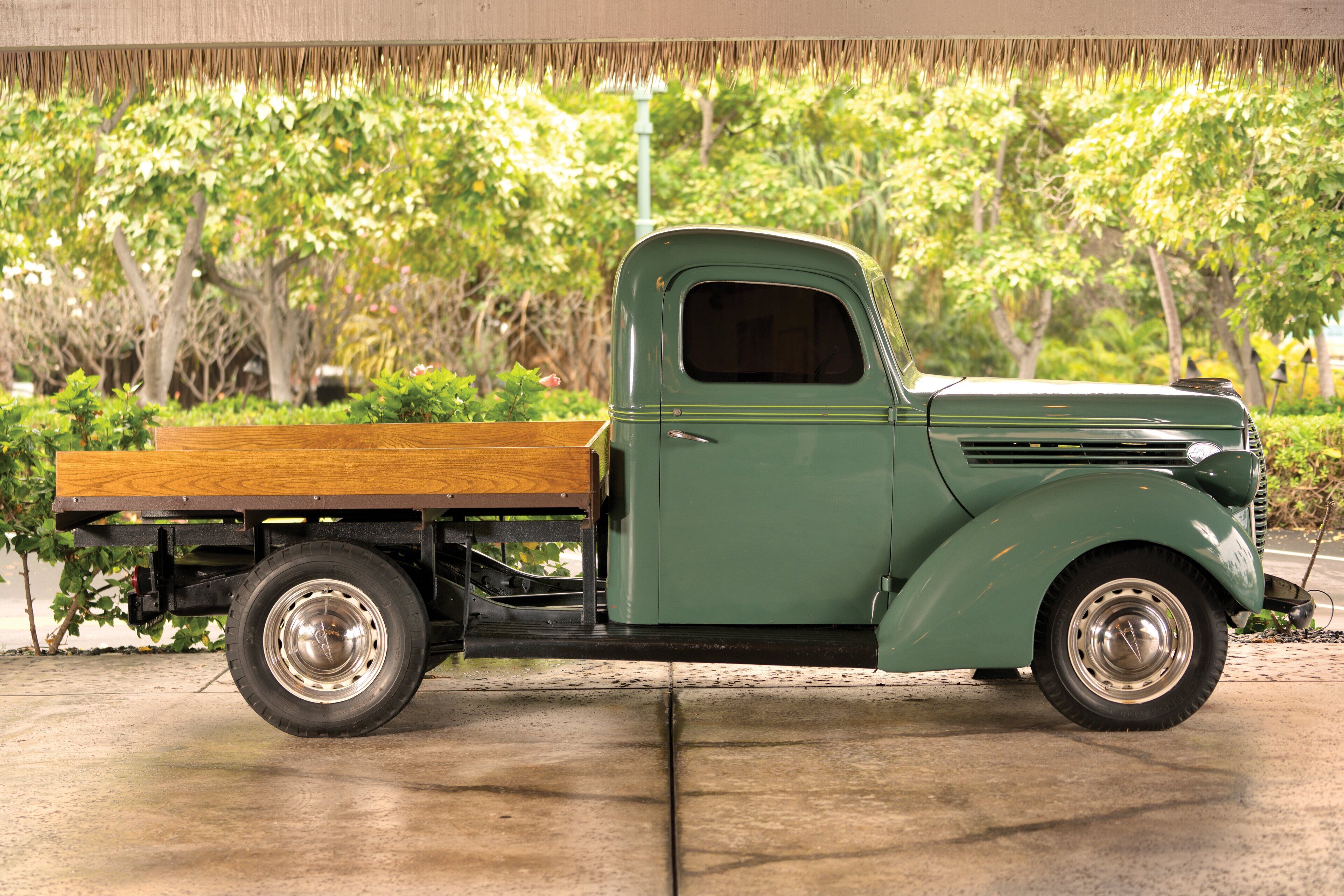 Una camioneta verde antigua estacionada sobre una superficie de concreto con caja de madera, rodeada de follaje.