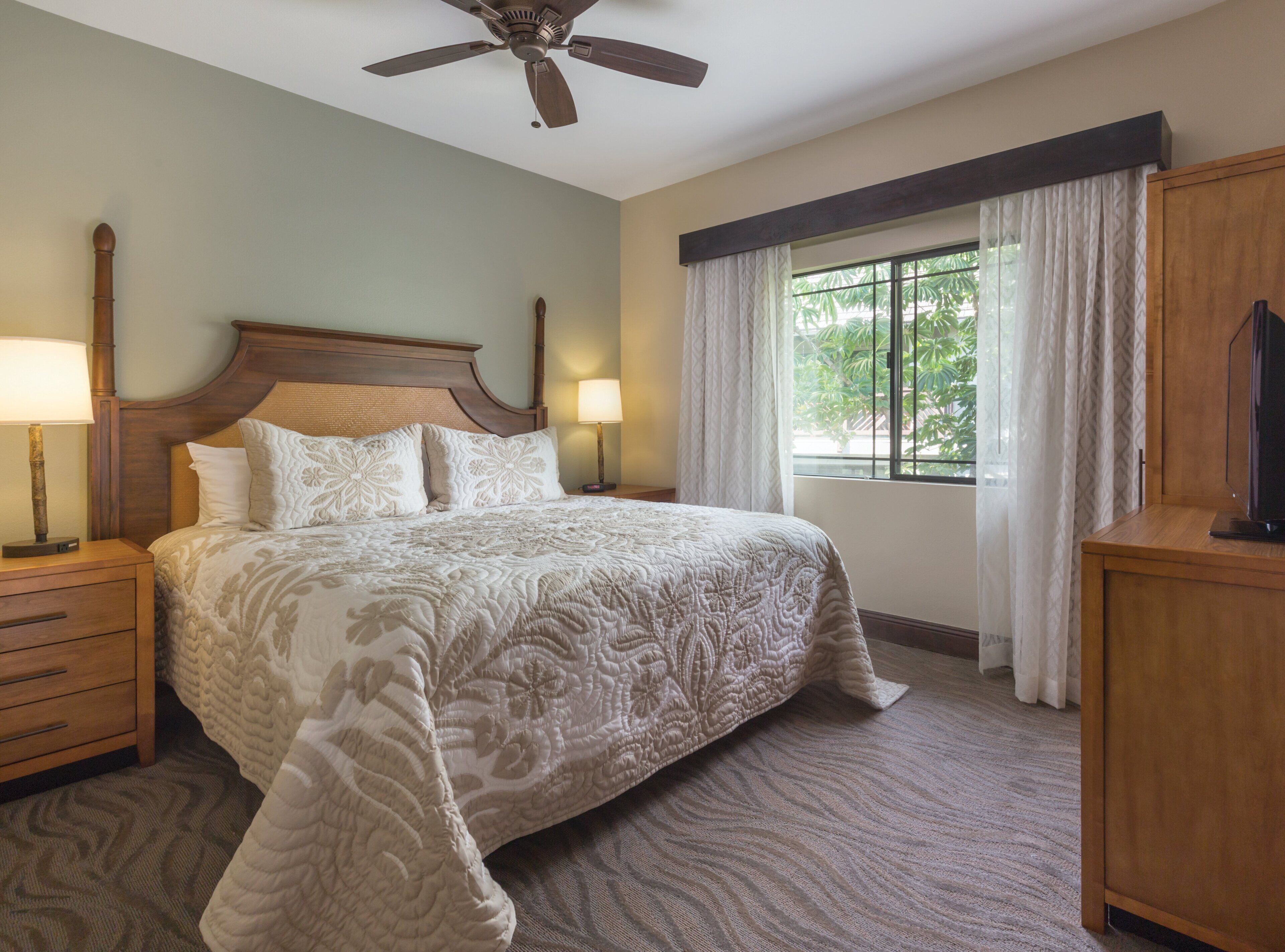 A hotel room with a large bed, a wooden headboard, a ceiling fan, a nightstand with a lamp, a wooden dresser, and a window with curtains.