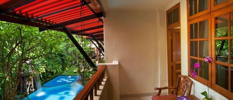 A balcony with a pool, wooden railing, and a view of lush greenery.