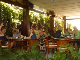 A group of people seated at tables in an outdoor cafe with wooden furniture and greenery in the background.