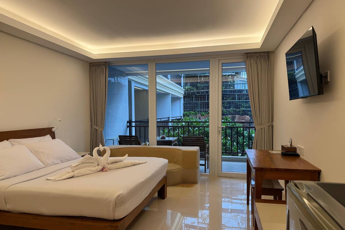 A hotel room with a bed, a desk, a chair, and a balcony with a view of greenery.