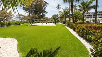 A well-maintained lawn with a volleyball net, surrounded by palm trees and a building in the background.