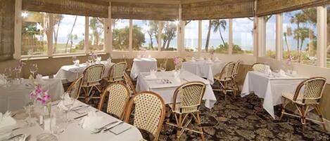Comedor con mesas dispuestas para el servicio, sillas de mimbre y ventanas amplias con vistas al océano.