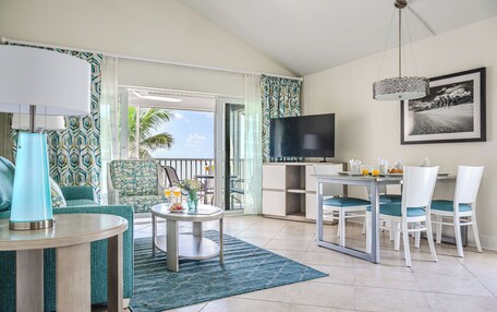 A modern living room with a TV, dining table, and chairs. There is a balcony with a view of palm trees.