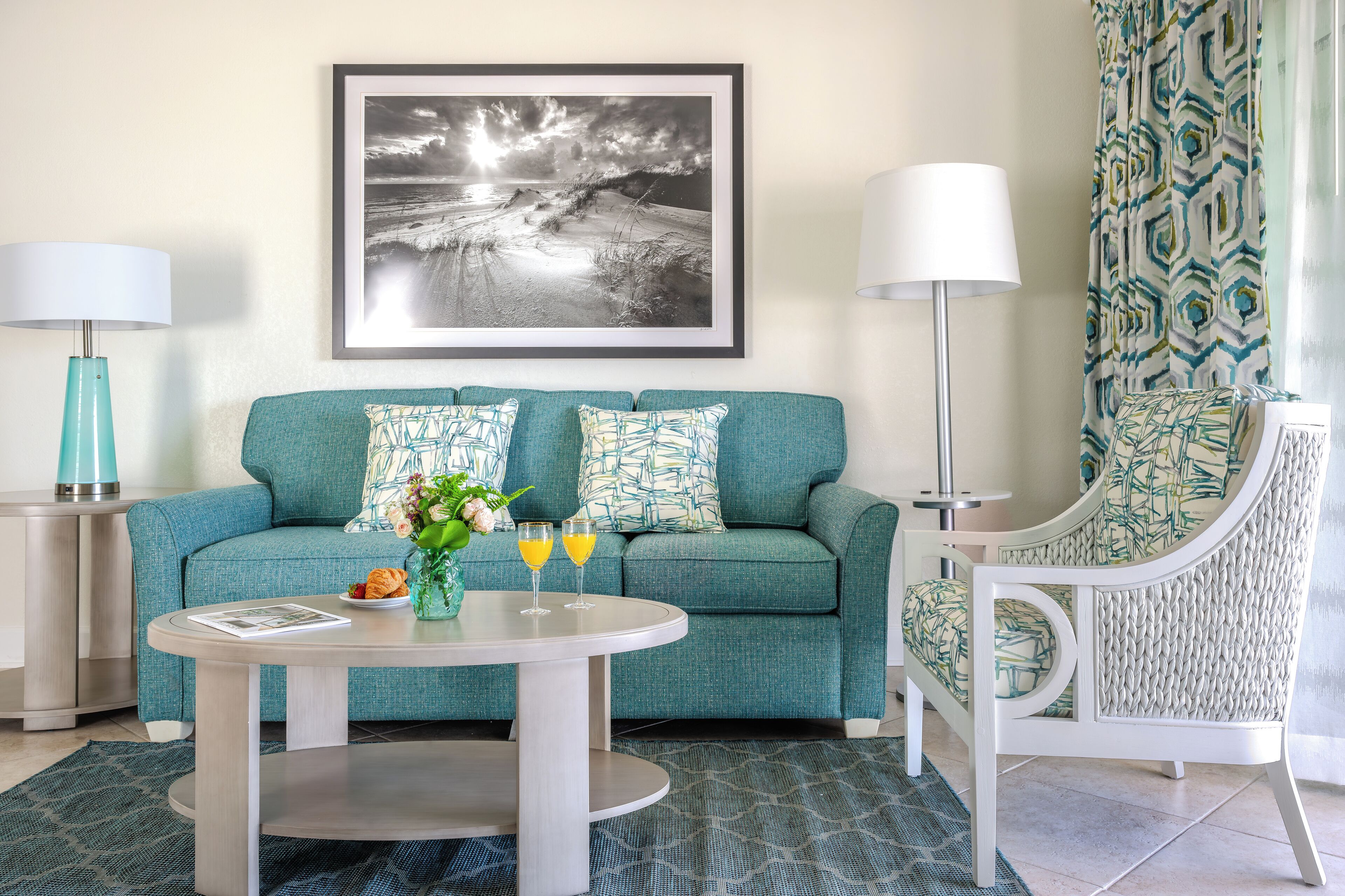 A teal sofa with patterned cushions, a round coffee table with a vase and two glasses, and a framed black and white landscape painting above.