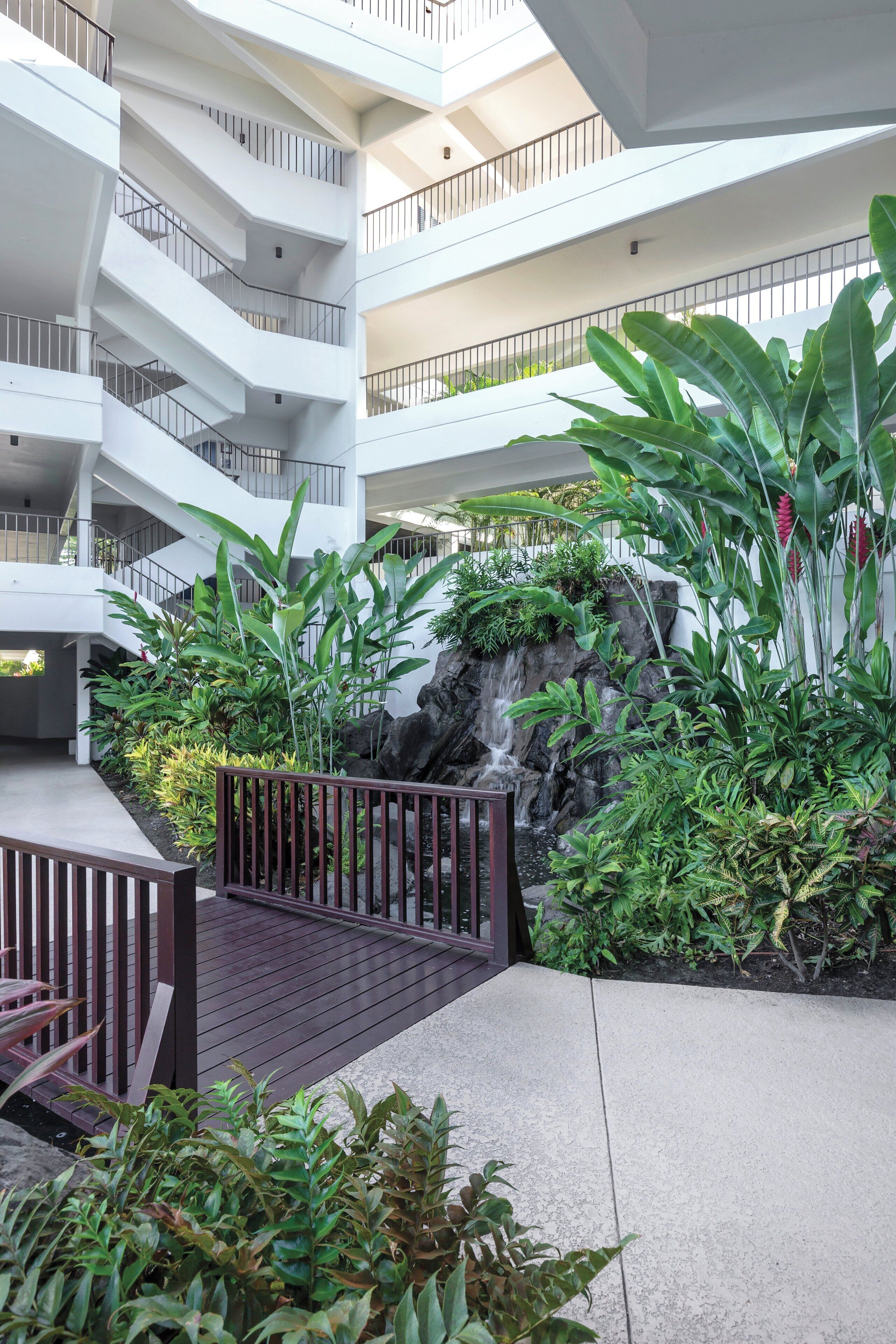 Ein mehrgeschossiges Atrium mit einer zentralen Wasserspiels, umgeben von üppiger Vegetation und einer Holzterrasse.