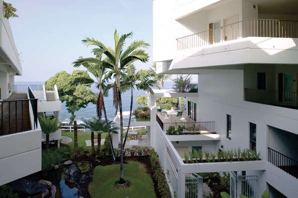 Un edificio residencial de varios pisos con balcones, un pequeño jardín con palmeras y vista al mar.