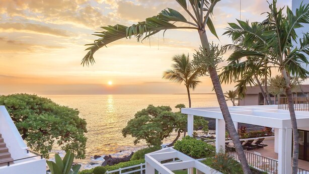 Een modern resort met uitzicht op zee bij zonsondergang, met een zwembad, palmbomen en een wit gebouw.