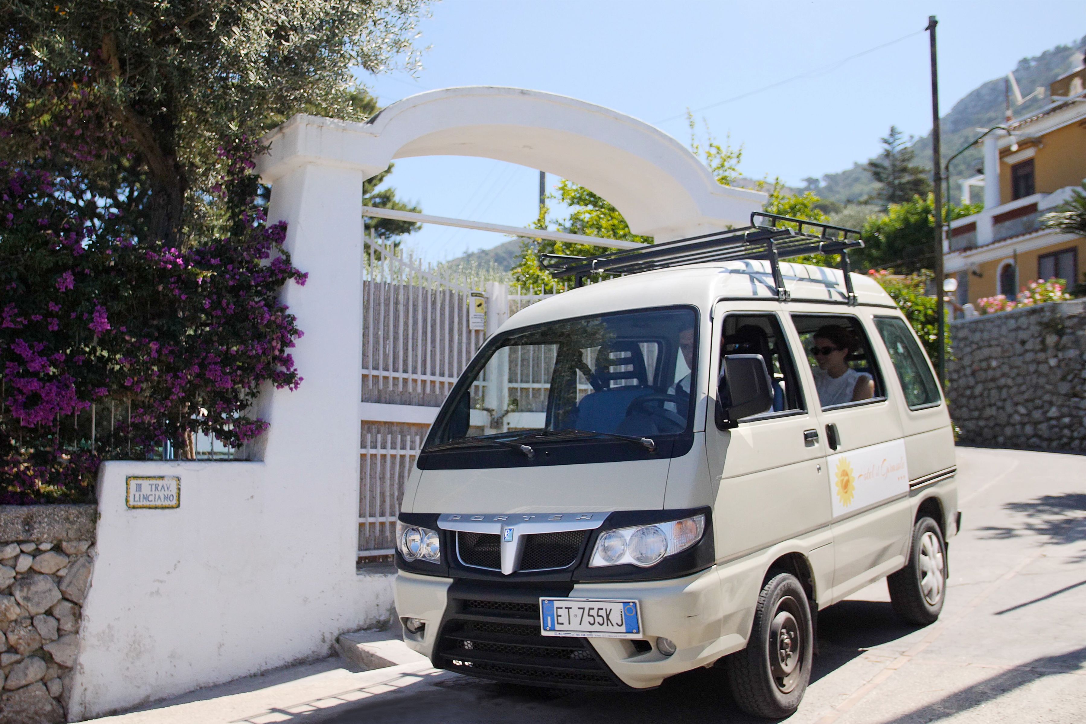 A white van parked in front of a white gate with a stone wall and a sign that reads "TRAV LINCIANO".