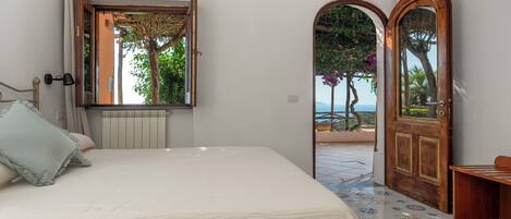 A bedroom with a large bed, a window with curtains, a radiator, and a view of a balcony with a tree.