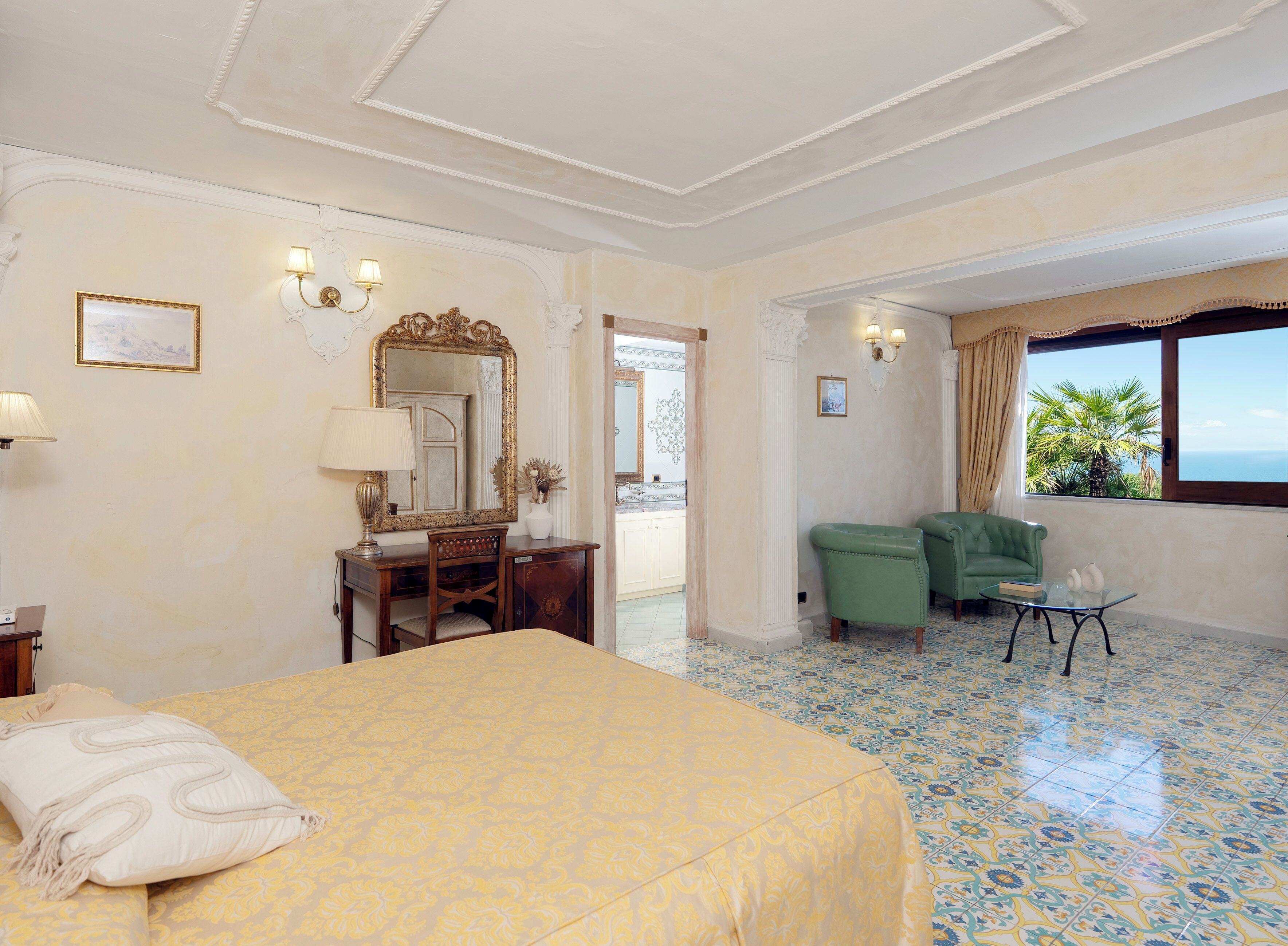 A hotel room with a bed, a desk, a green armchair, a window with a view, and a tiled floor.