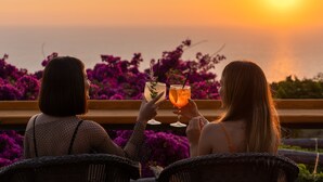 Two people sitting on wicker chairs facing a sunset over the sea, holding drinks. - Hotel Il Girasole (Anacapri)