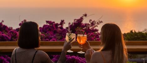 Two people sitting on wicker chairs facing a sunset over the sea, holding drinks.