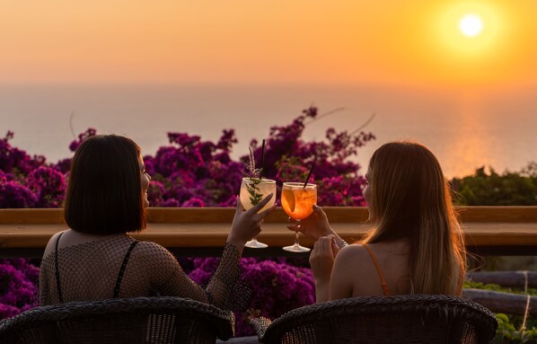 Deux personnes sont assises sur des chaises en osier, face au soleil couchant sur la mer, et tiennent chacune un verre à la main.