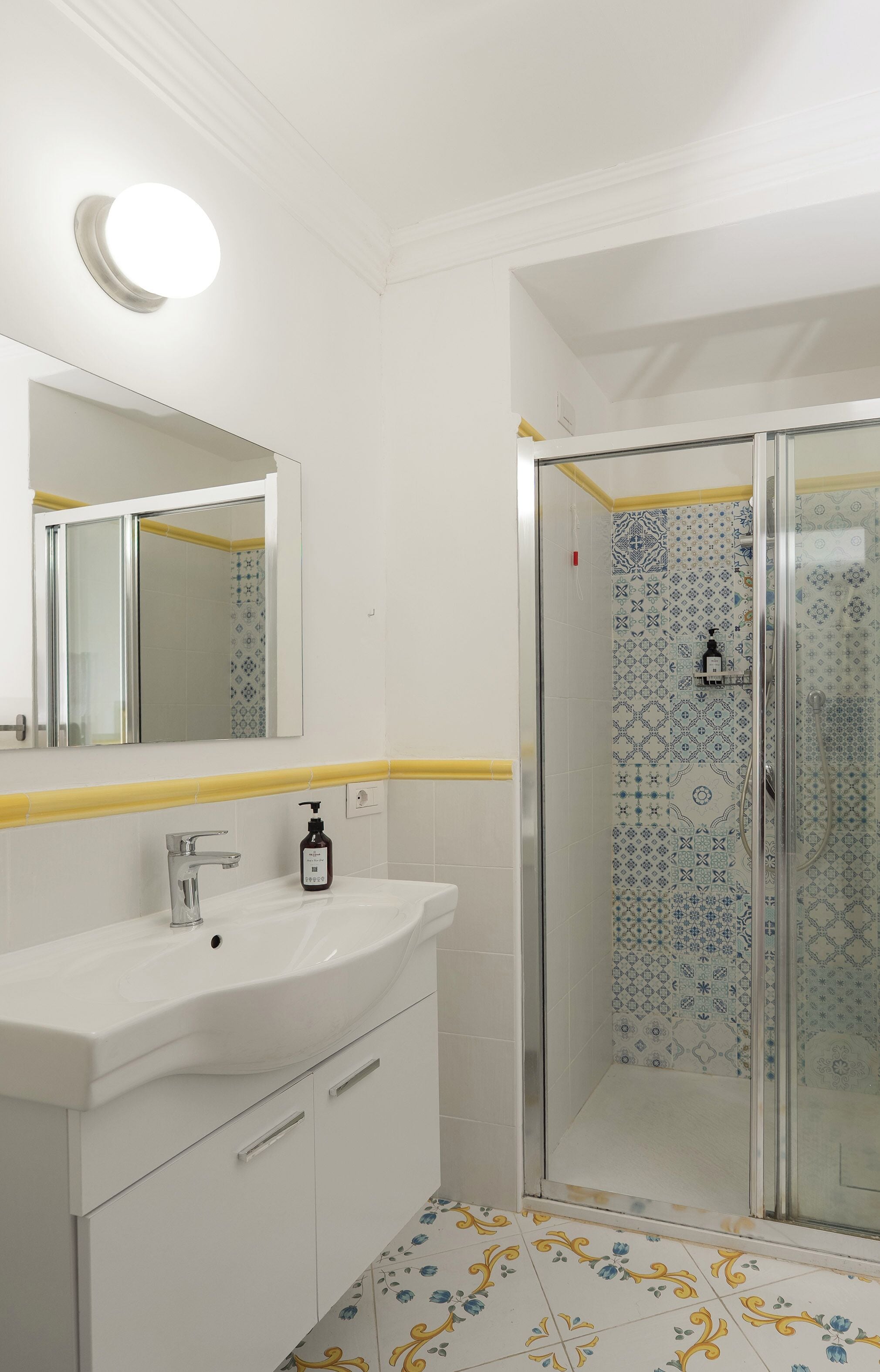 A bathroom with a white sink, mirror, and yellow-tiled floor. It includes a shower with patterned tiles and a glass door.