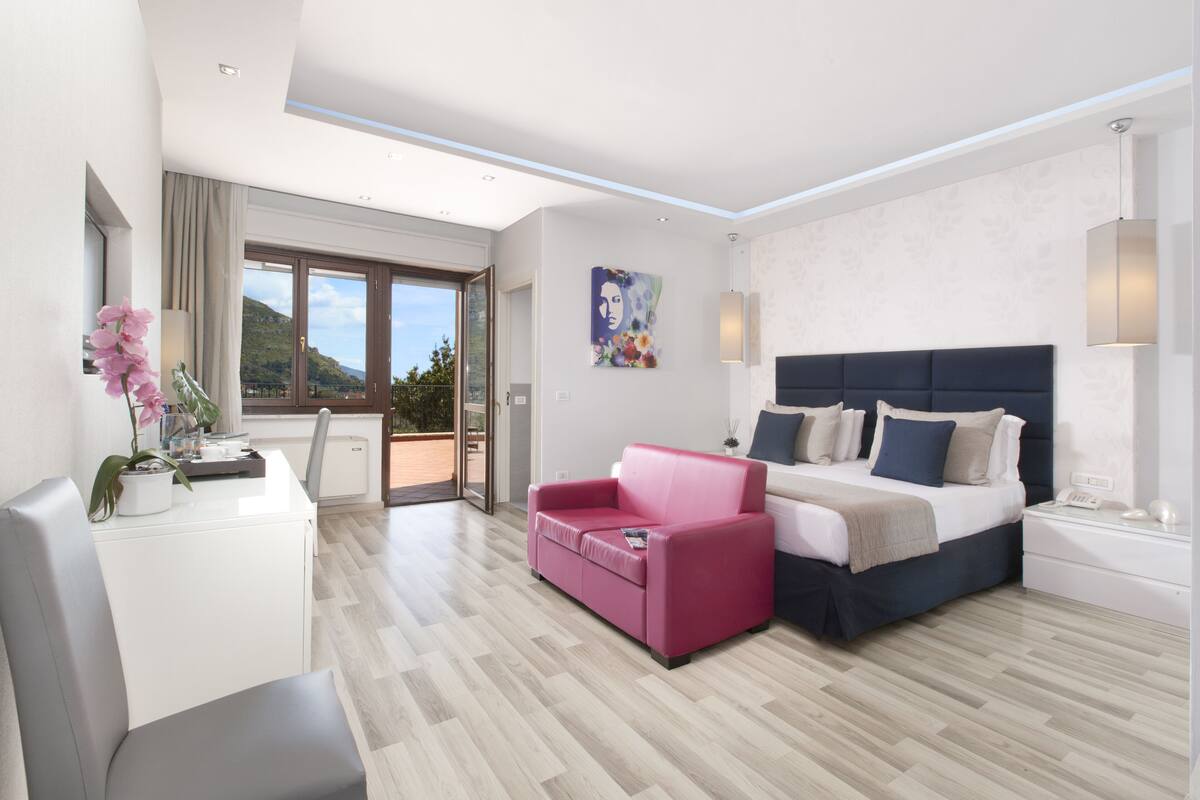 A modern hotel room with a bed, a pink sofa, a desk, and a view of the outdoors.