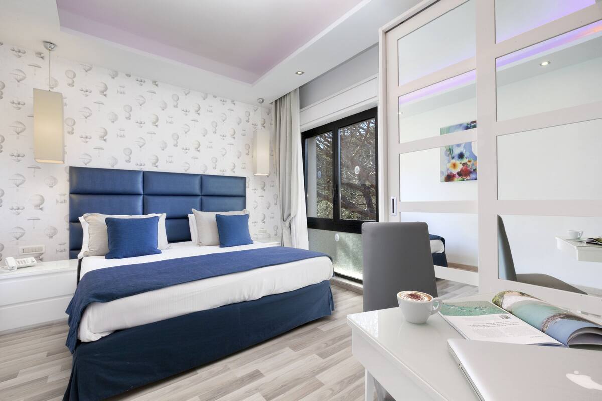 A modern bedroom with a large bed, a desk with a laptop, and a window with a view of trees.